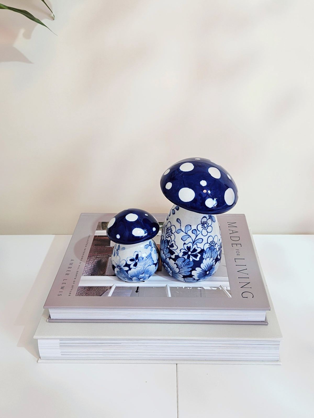 Two ceramic mushroom-shaped decorative objects in a blue and white floral and polka-dot pattern, displayed on top of stacked books against a light beige wall.