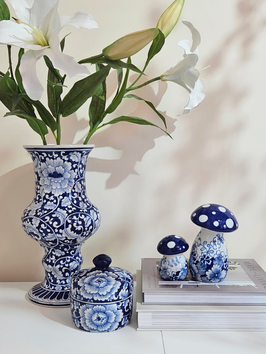 Blue and white floral porcelain ceramic set including a tall vase with white lilies, a small lidded box, and two mushroom-shaped decor pieces on stacked books against a warm beige wall.
