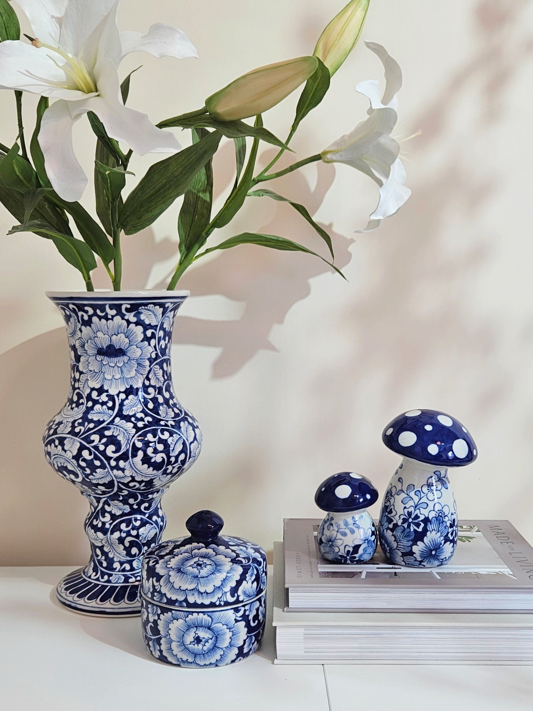 Blue and white floral porcelain ceramic set including a tall vase with white lilies, a small lidded box, and two mushroom-shaped decor pieces on stacked books against a warm beige wall.