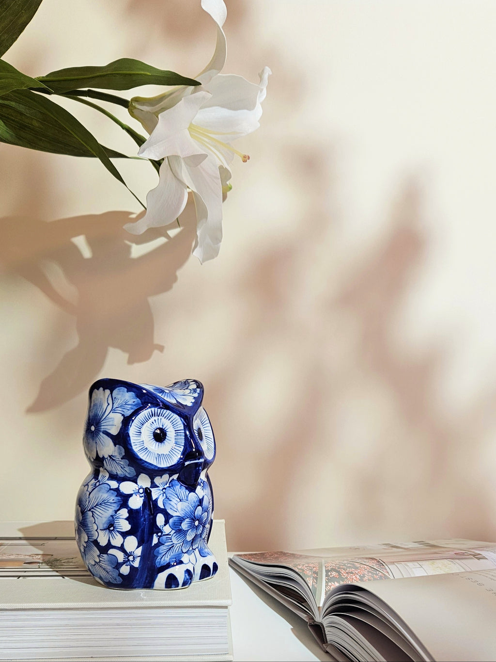 A small ceramic owl figurine in a cobalt blue and white floral pattern sits on a white surface next to an open book, with white lilies and soft shadows in the background.