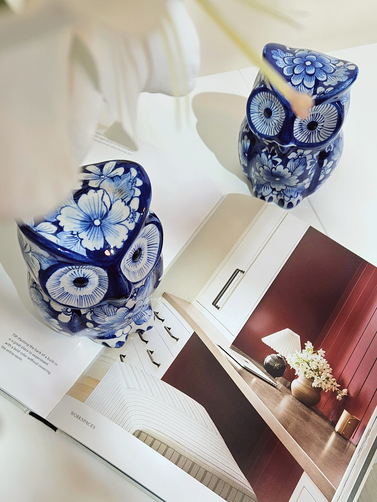 Two blue and white chinoiserie floral owl figurines displayed on an open magazine, with a white flower near one owl.
