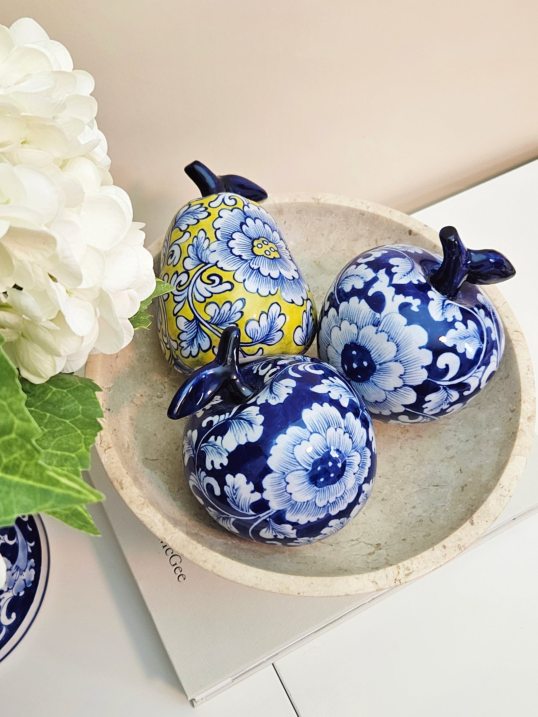 Three decorative ceramic apples and  pears figurines with floral patterns in blue, white, and yellow, chinoiserie style, arranged in a shallow beige bowl on a white surface next to white flowers.