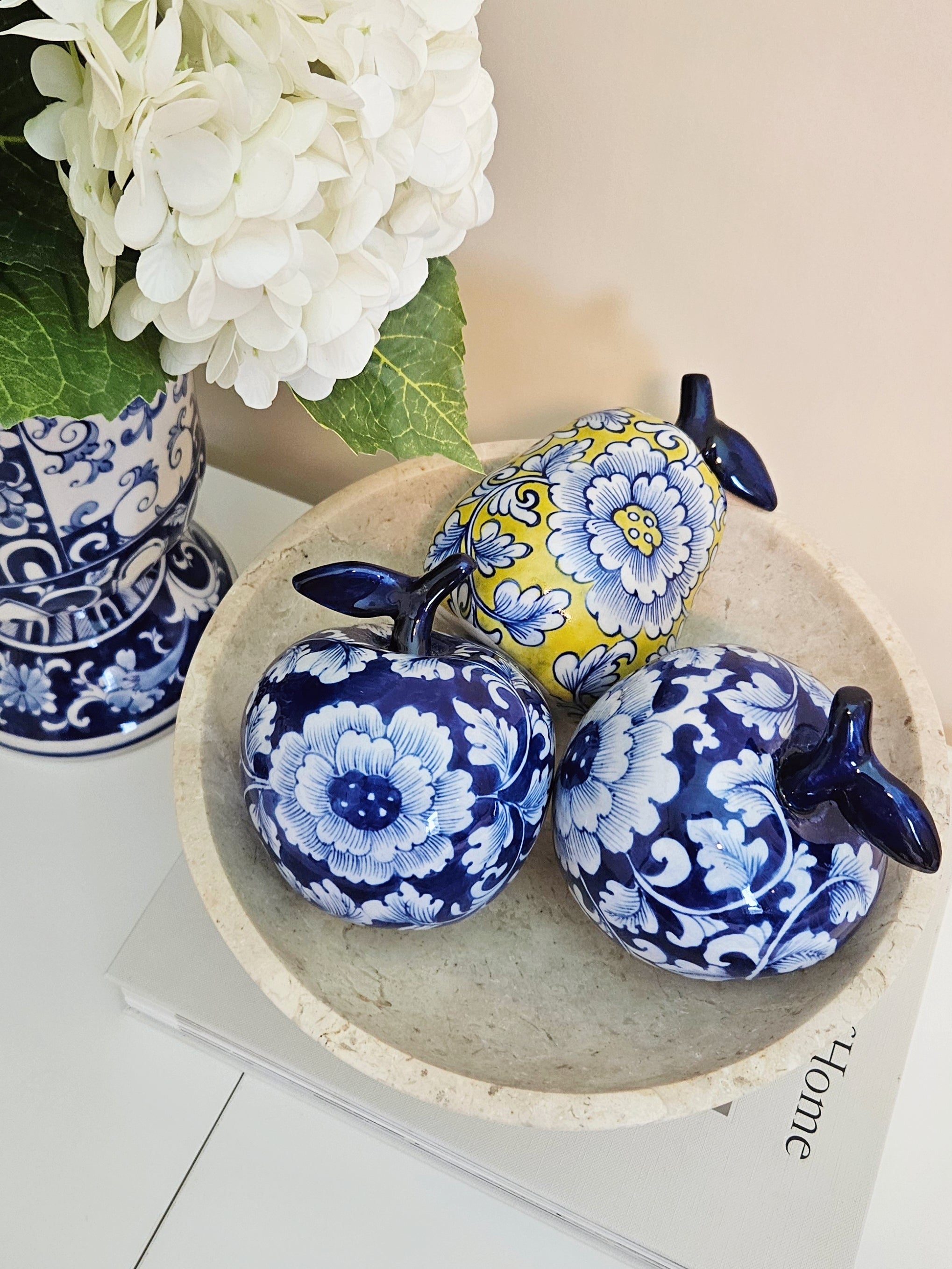 two decorative ceramic apples with blue and white floral patterns and one yellow pear, arranged in a round stone tray on a tabletop next to a blue and white patterned vase with white flowers.