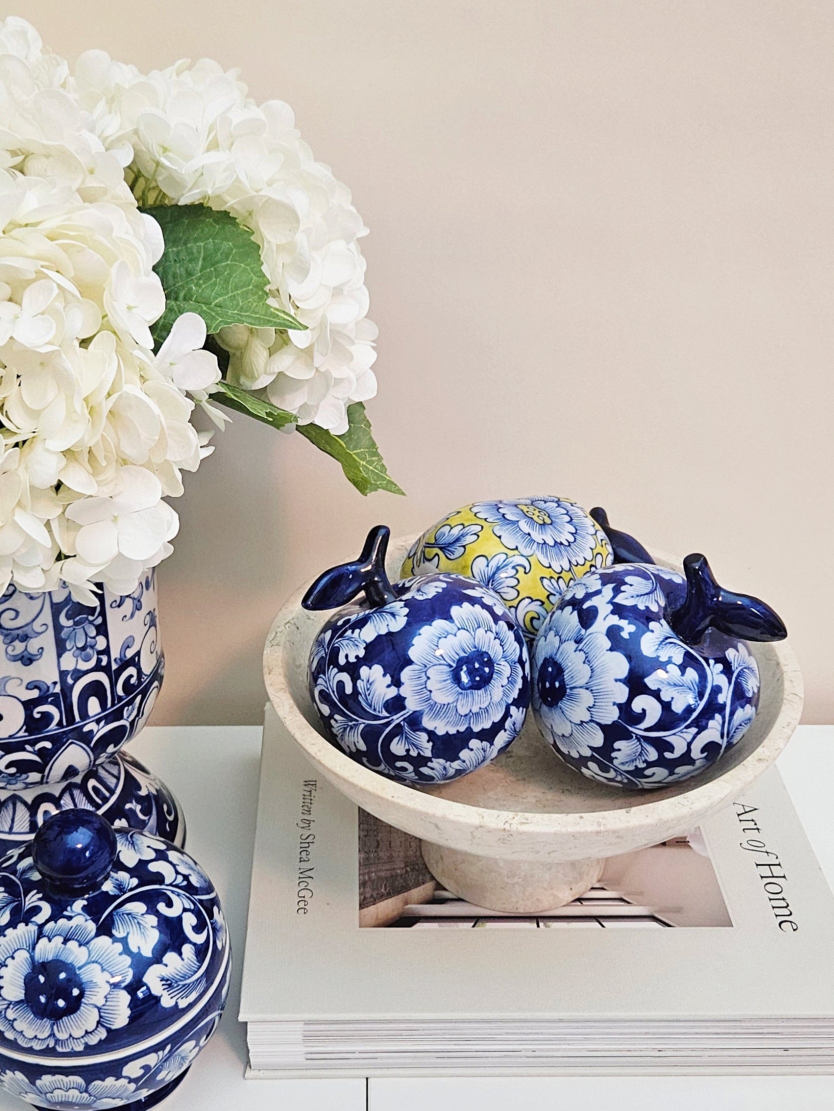 two decorative ceramic apples in blue and white floral patterns with one yellow patterned pear, displayed on a pedestal bowl on top of a magazine next to a blue and white vase of white flowers.