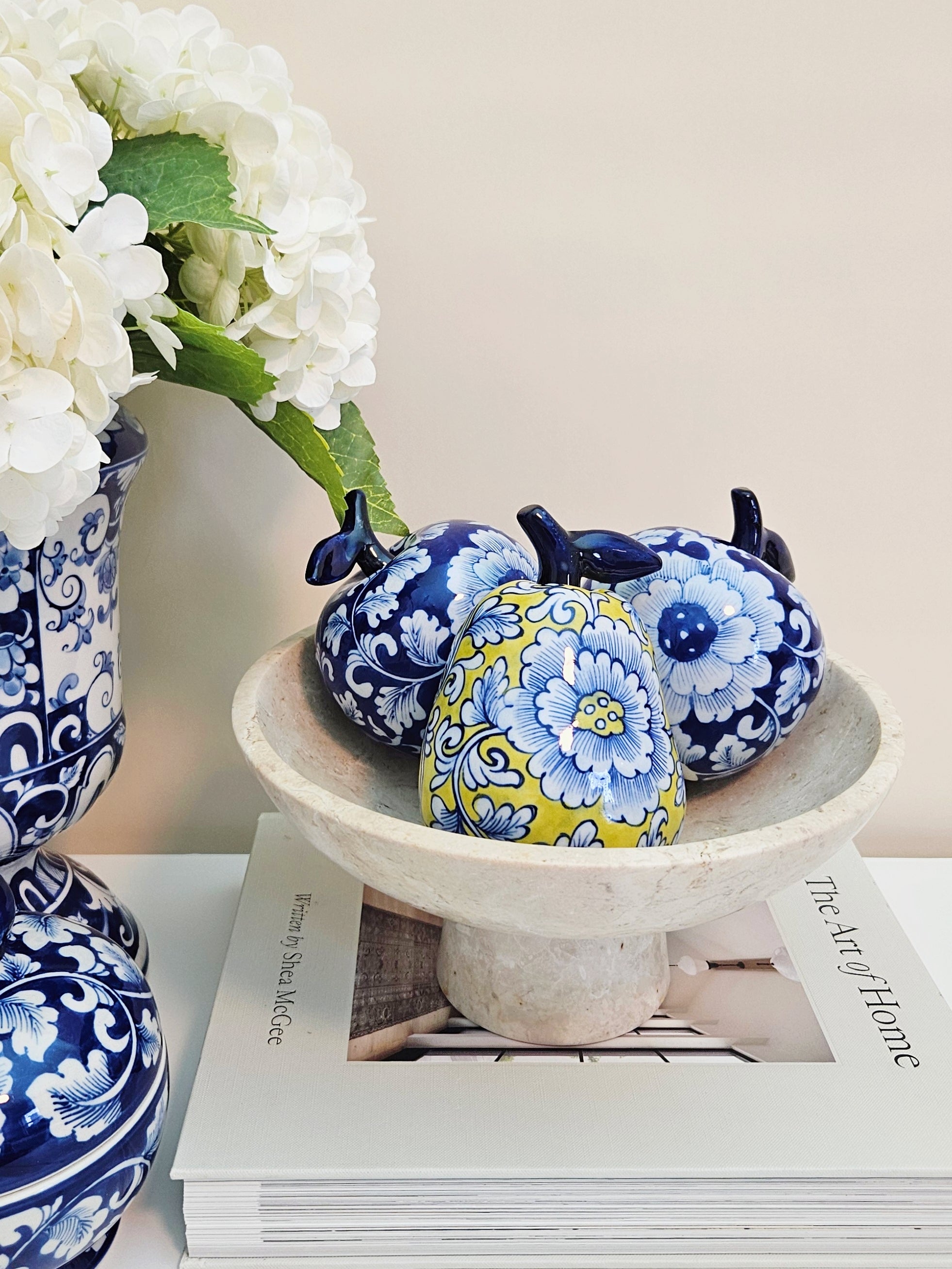 Three decorative ceramic apples and pear with blue and yellow floral patterns are arranged in a white pedestal bowl on top of a magazine, next to a blue and white floral vase with white flowers.