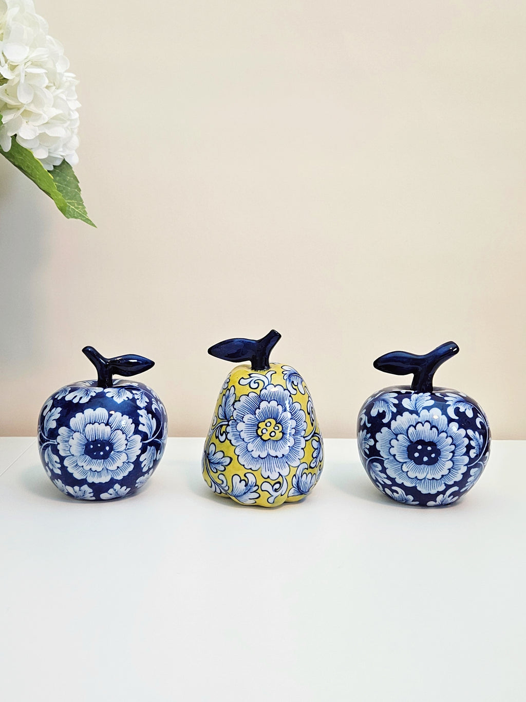 Three decorative ceramic fruit figurines with floral patterns, including two blue-and-white apples and one yellow-and-blue pear, displayed on a white surface against a beige background.