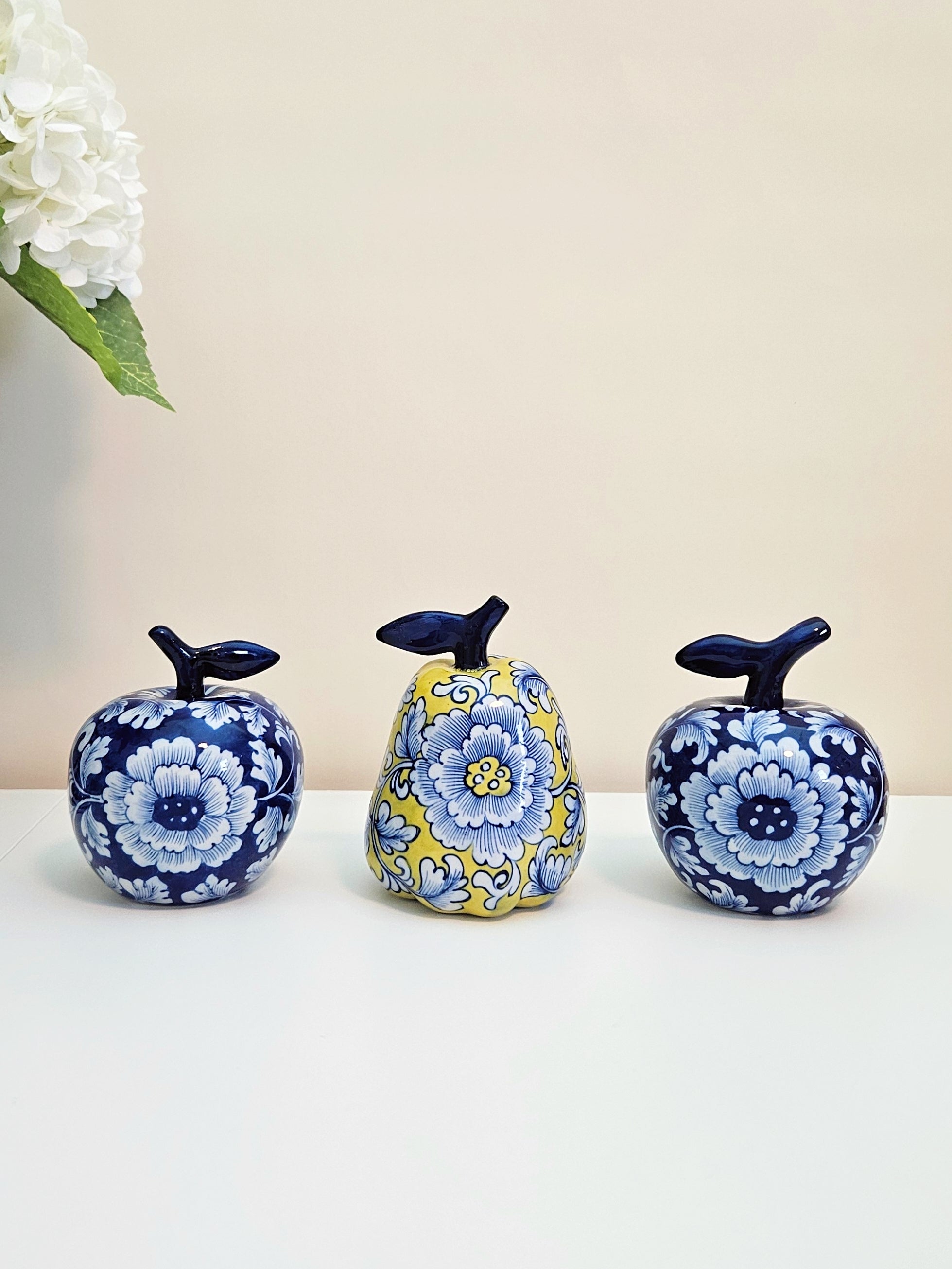Three decorative ceramic fruit figurines with floral patterns, including two blue-and-white apples and one yellow-and-blue pear, displayed on a white surface against a beige background.