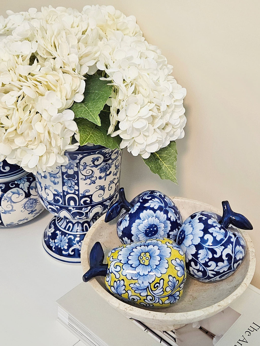 Decorative ceramic fruit-shaped figurines with blue and yellow floral patterns arranged in a white bowl next to a blue and white floral vase filled with white flowers.