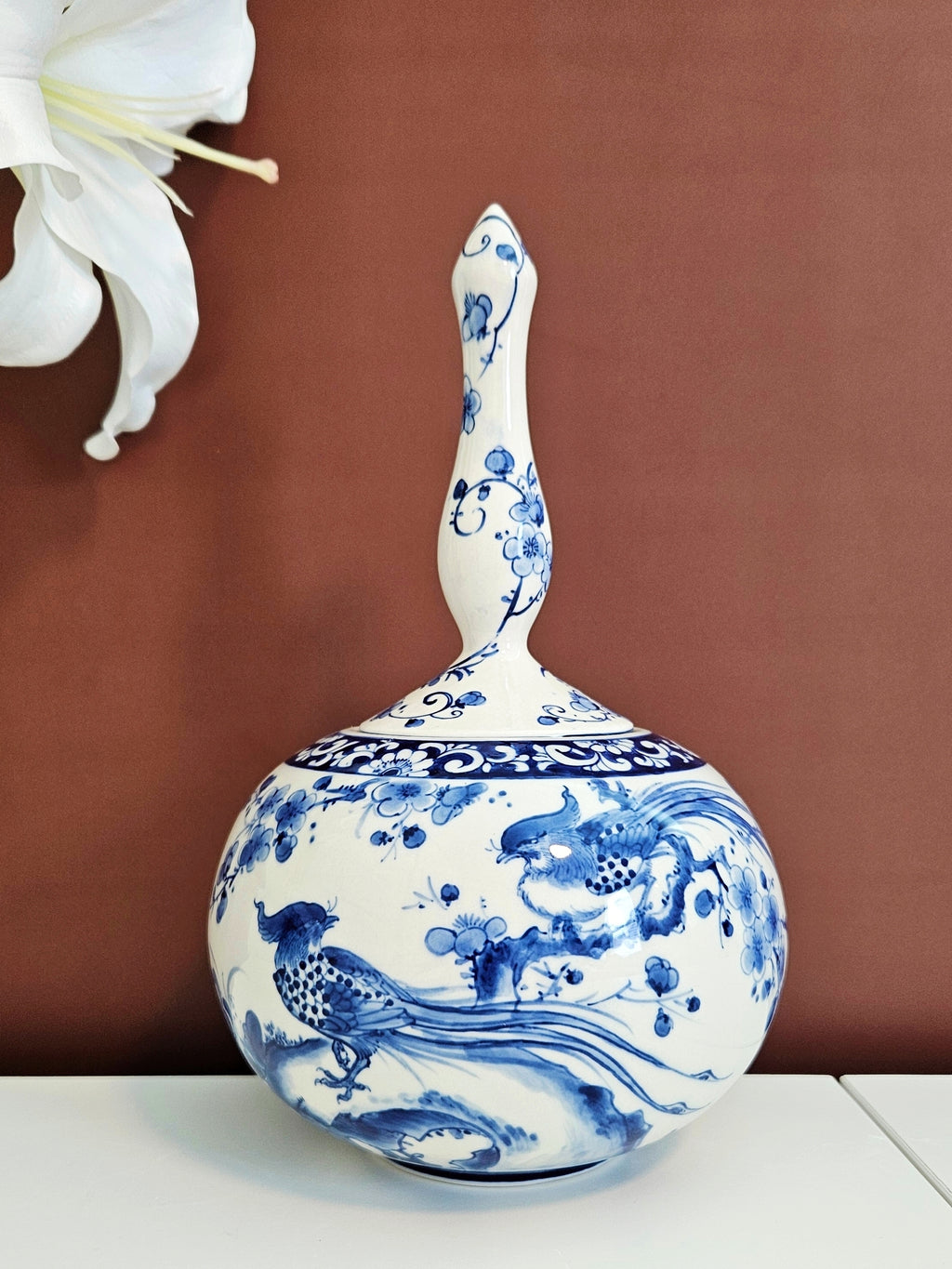 A ceramic round-shaped vase with a unique tall lid, decorated in traditional blue and white porcelain chinoiserie style with a pheasant motif, set against a warm brown background.