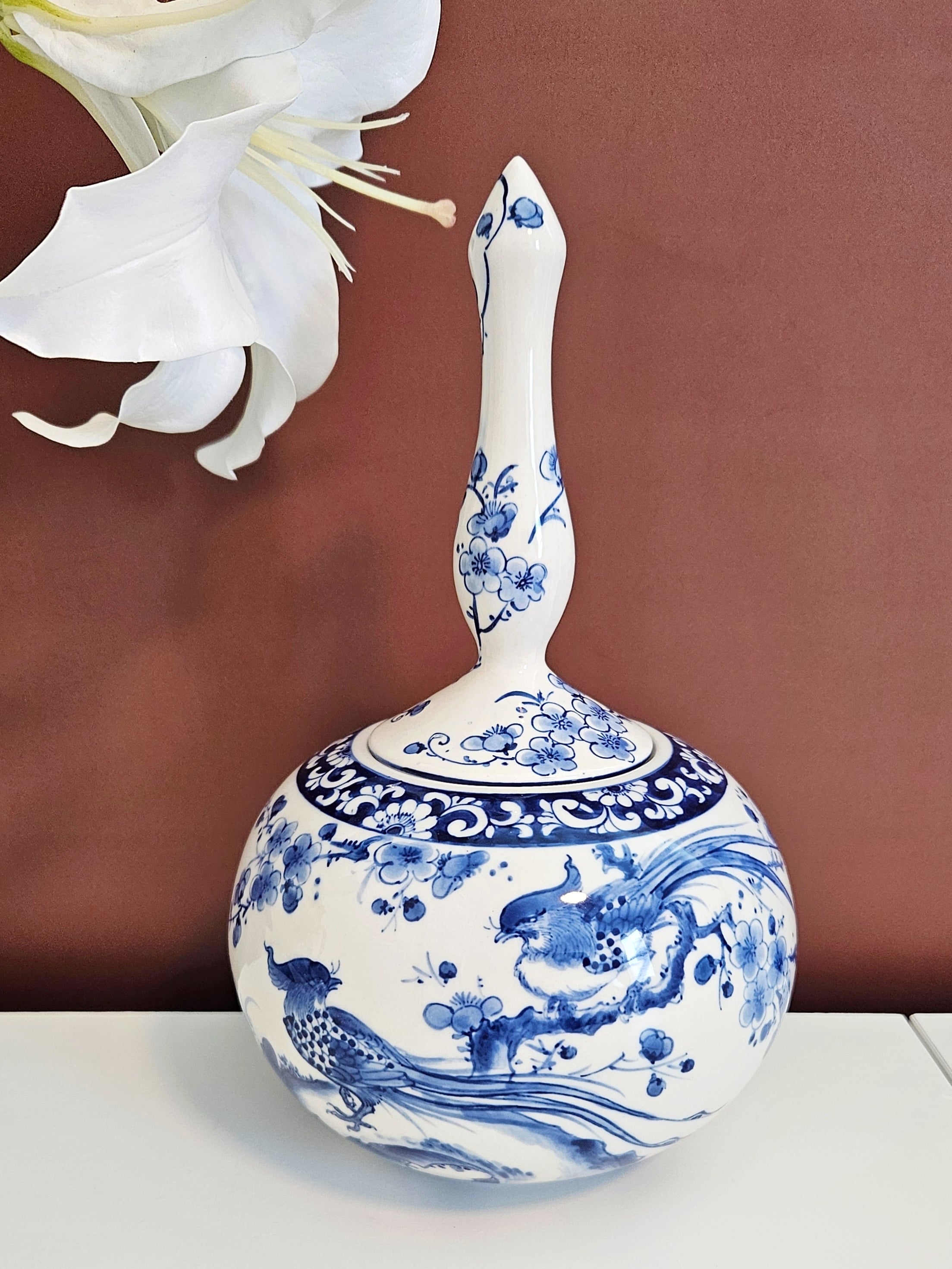 A ceramic round-shaped jar with a blue and white hand-painted floral and pheasant pattern, set against a solid brown background with a white lily flower in the upper left corner.