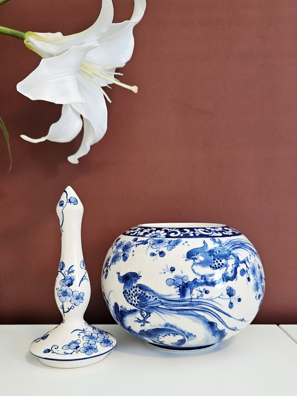 A blue and white chinoiserie porcelain set consisting of a round canister and a matching tall lid, displayed on a white surface against a brown background with a white lily flower in the upper left corner.