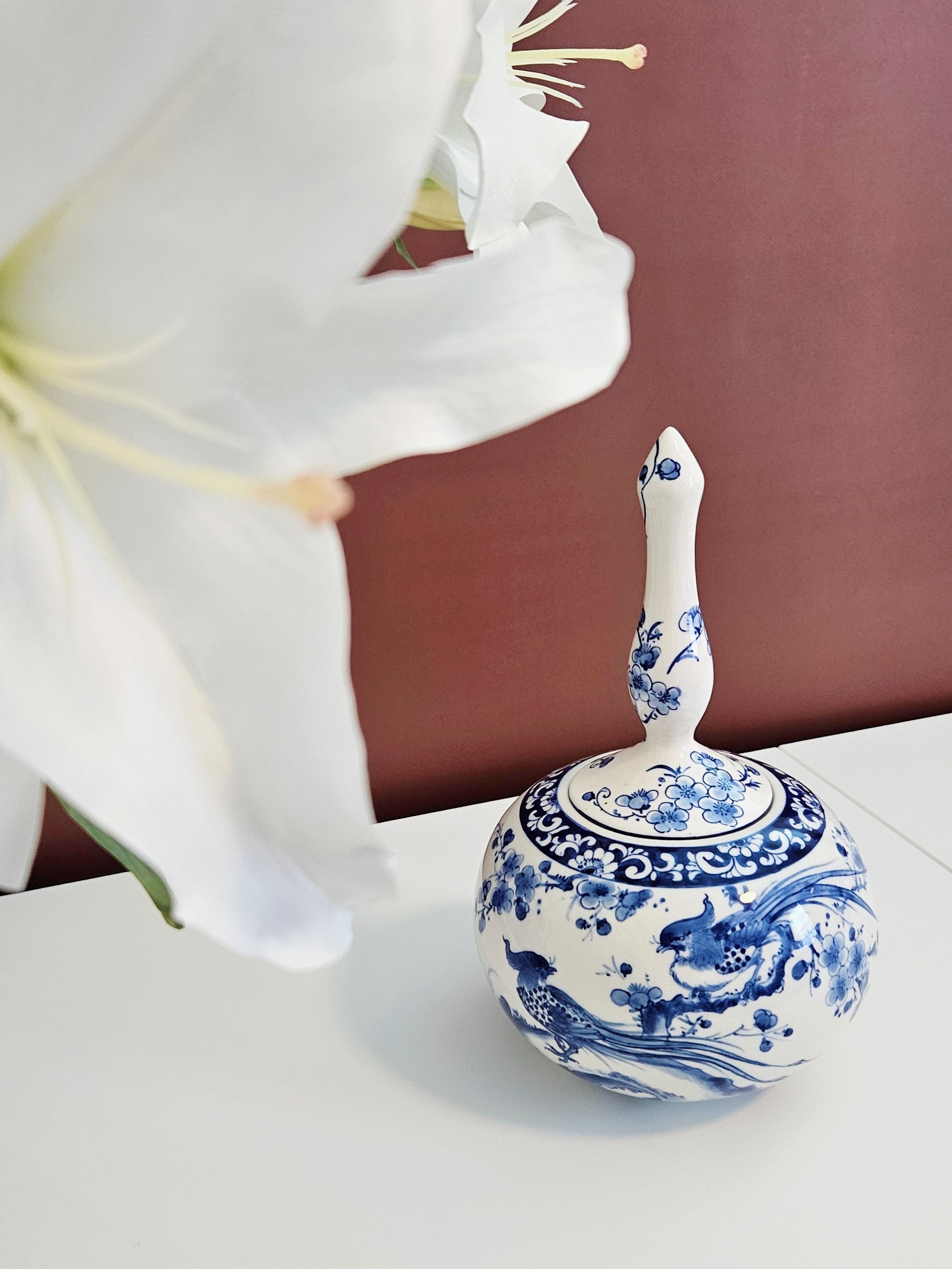 A small blue and white ceramic decorative jar with blue floral and pheasant patterns, featuring a narrow neck and tall lid, placed on a white surface against a dark reddish-brown background, with a large white flower partially framing the left side of the shot.
