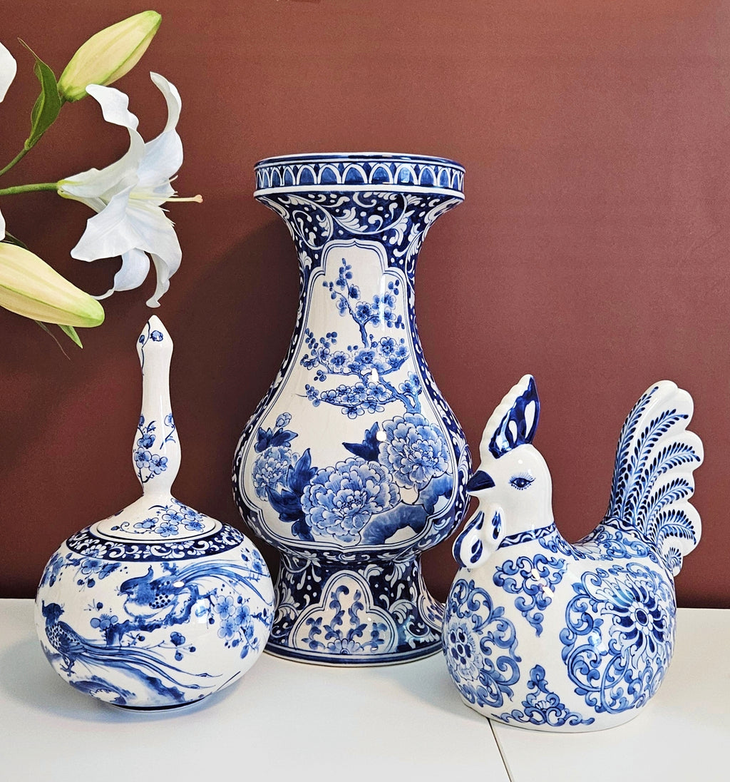Three blue and white porcelain decorative pieces with traditional floral and wave patterns, arranged on a white surface against a warm reddish-brown background. One tall vase stands in the center, a smaller lidded jar is on the left, and a rooster figurine is on the right.