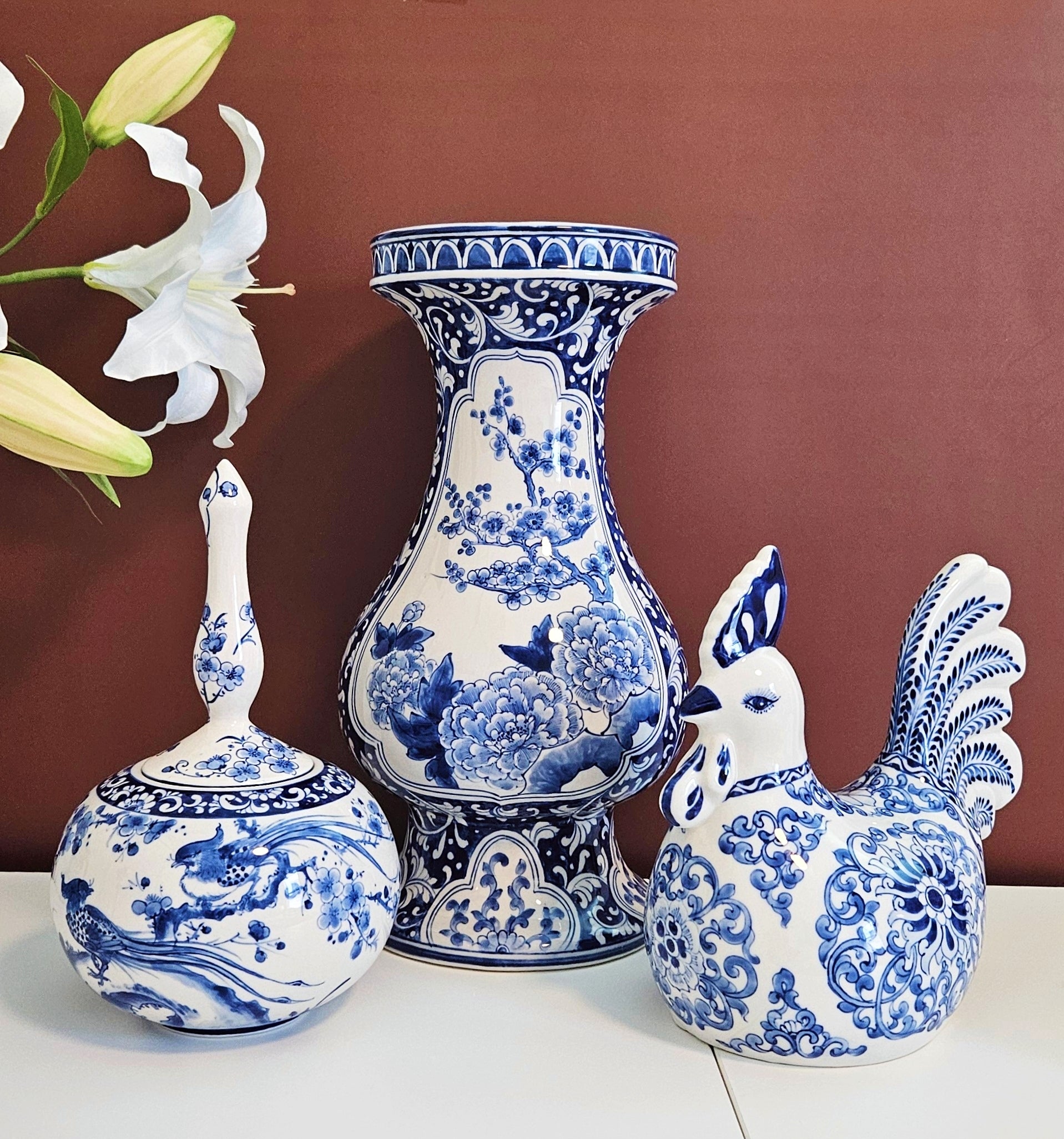 Three blue and white porcelain decorative pieces with traditional floral and wave patterns, arranged on a white surface against a warm reddish-brown background. One tall vase stands in the center, a smaller lidded jar is on the left, and a rooster figurine is on the right.