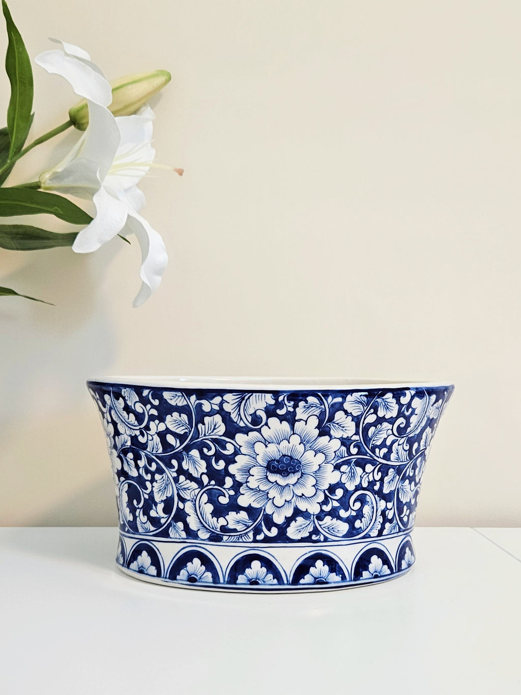 A blue and white chinoiserie porcelain planter bowl with an intricate floral pattern, displayed on a white surface against a beige wall with white lilies in the background.