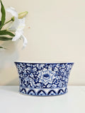 A blue and white chinoiserie porcelain planter bowl with an intricate floral pattern, displayed on a white surface against a beige wall with white lilies in the background.