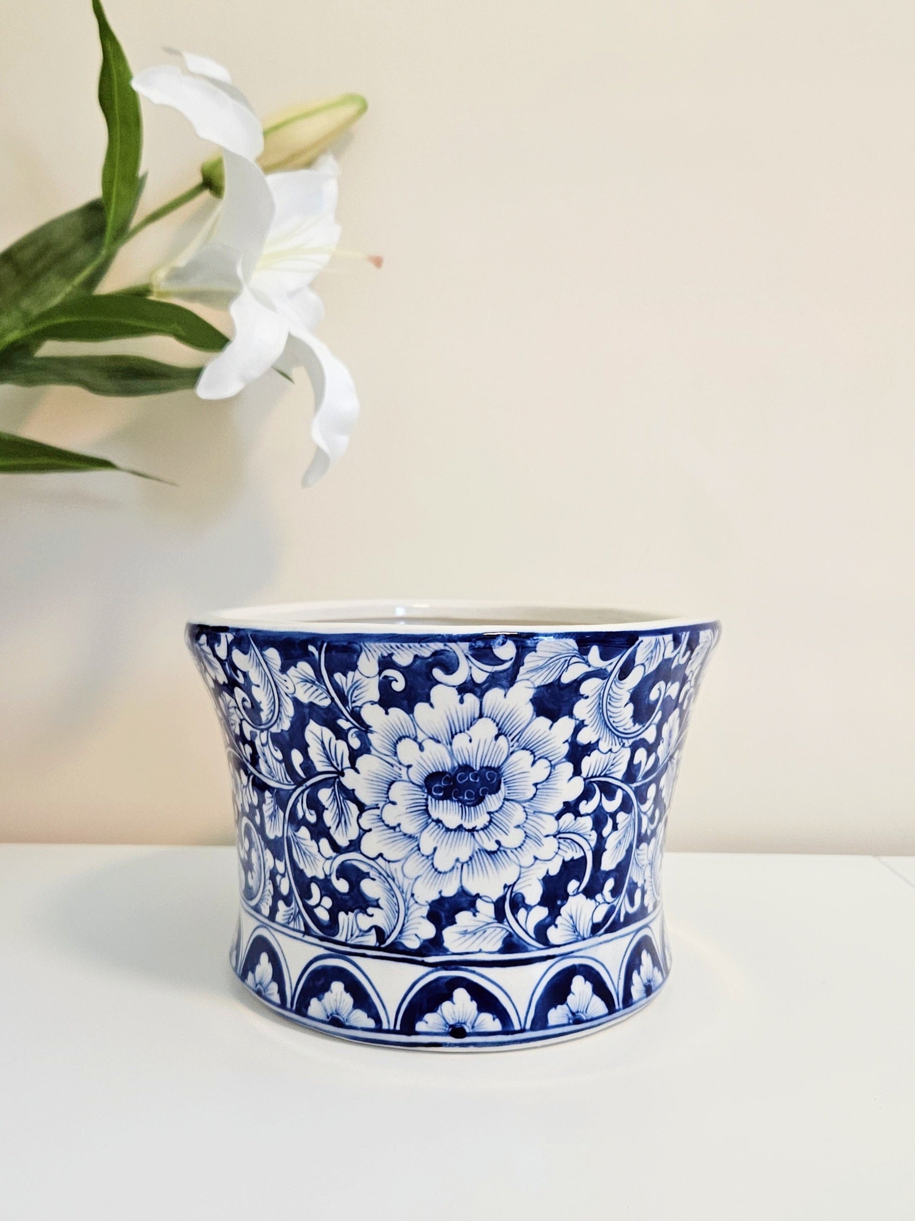 A blue and white porcelain planter pot with a floral pattern, chinoiserie style, displayed on a white surface against a light beige background with a white lily flower in the upper left corner.