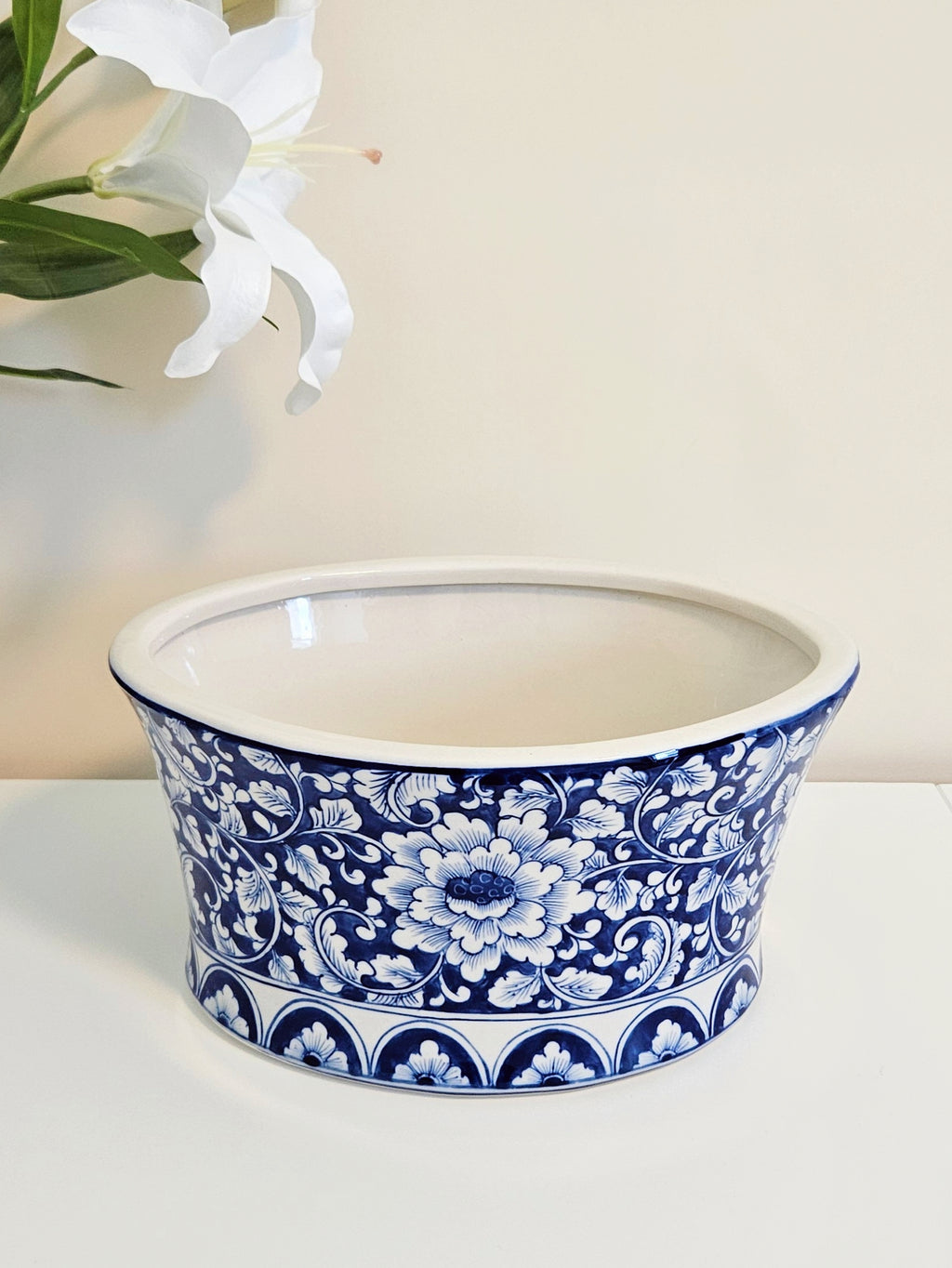 A round ceramic planter or bowl with a blue and white floral pattern, set on a white surface against a light beige background with a white lily flower in the upper left corner.