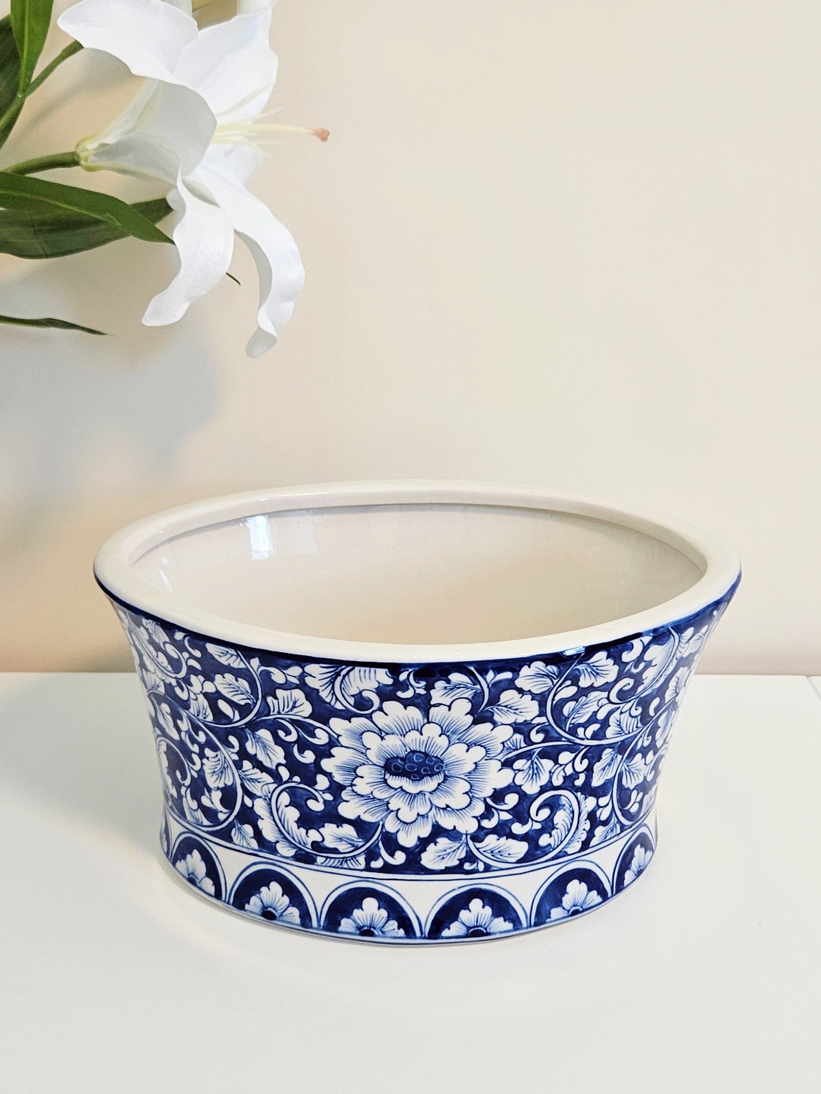 A round ceramic planter or bowl with a blue and white floral pattern, set on a white surface against a light beige background with a white lily flower in the upper left corner.