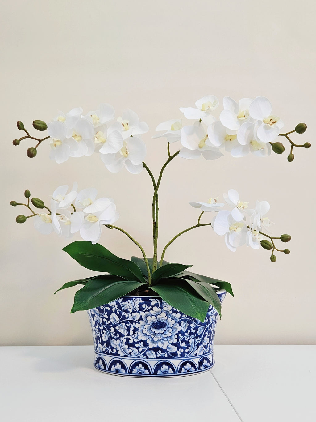 White artificial orchid flowers with green stems and leaves, arranged in a blue and white floral patterned ceramic pot on a white surface against a beige background.