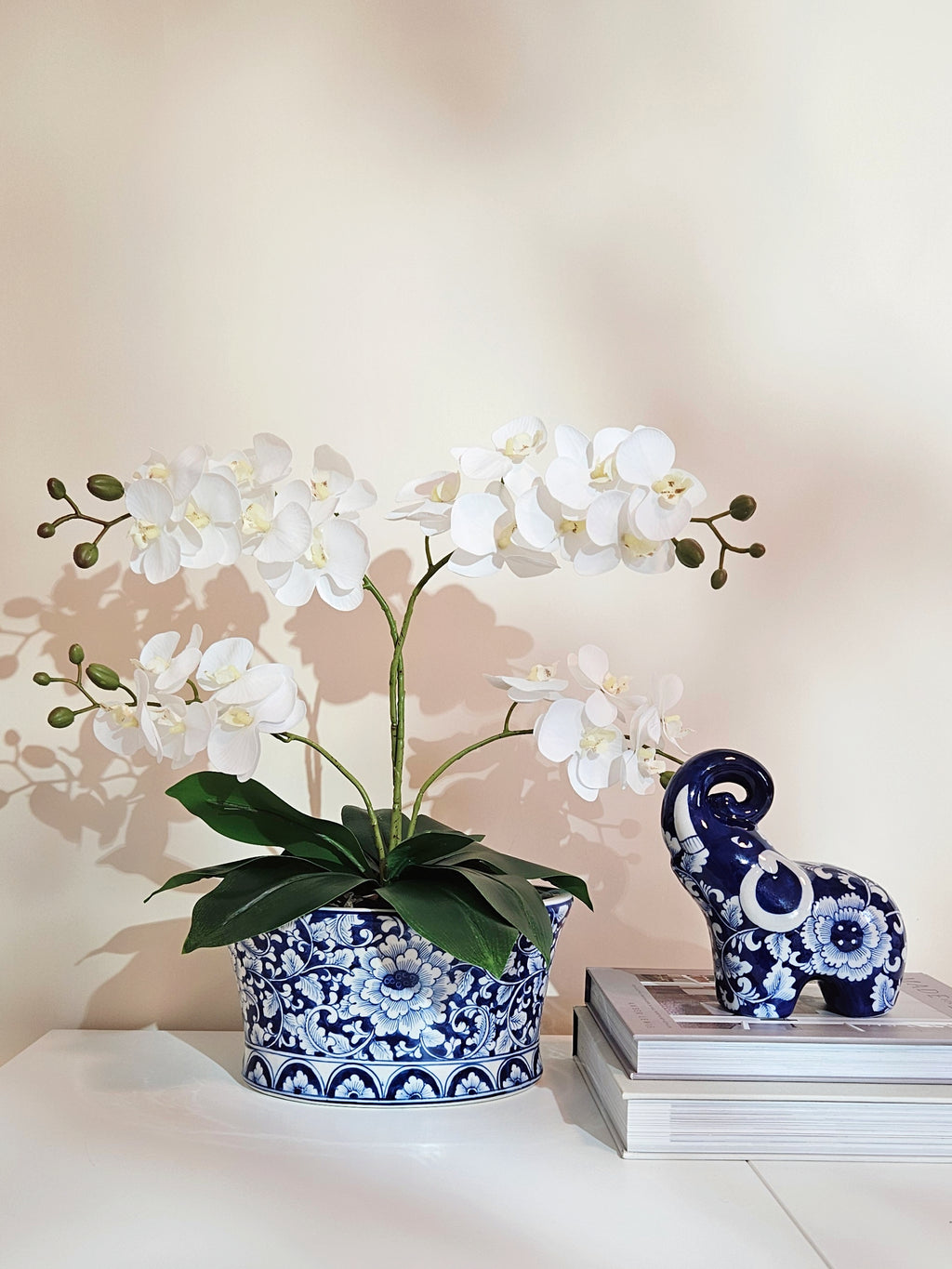 White artificial orchid flowers in a blue and white patterned ceramic planter pot, styled next to a matching blue and white elephant figurine on a stack of books against a warm beige wall.