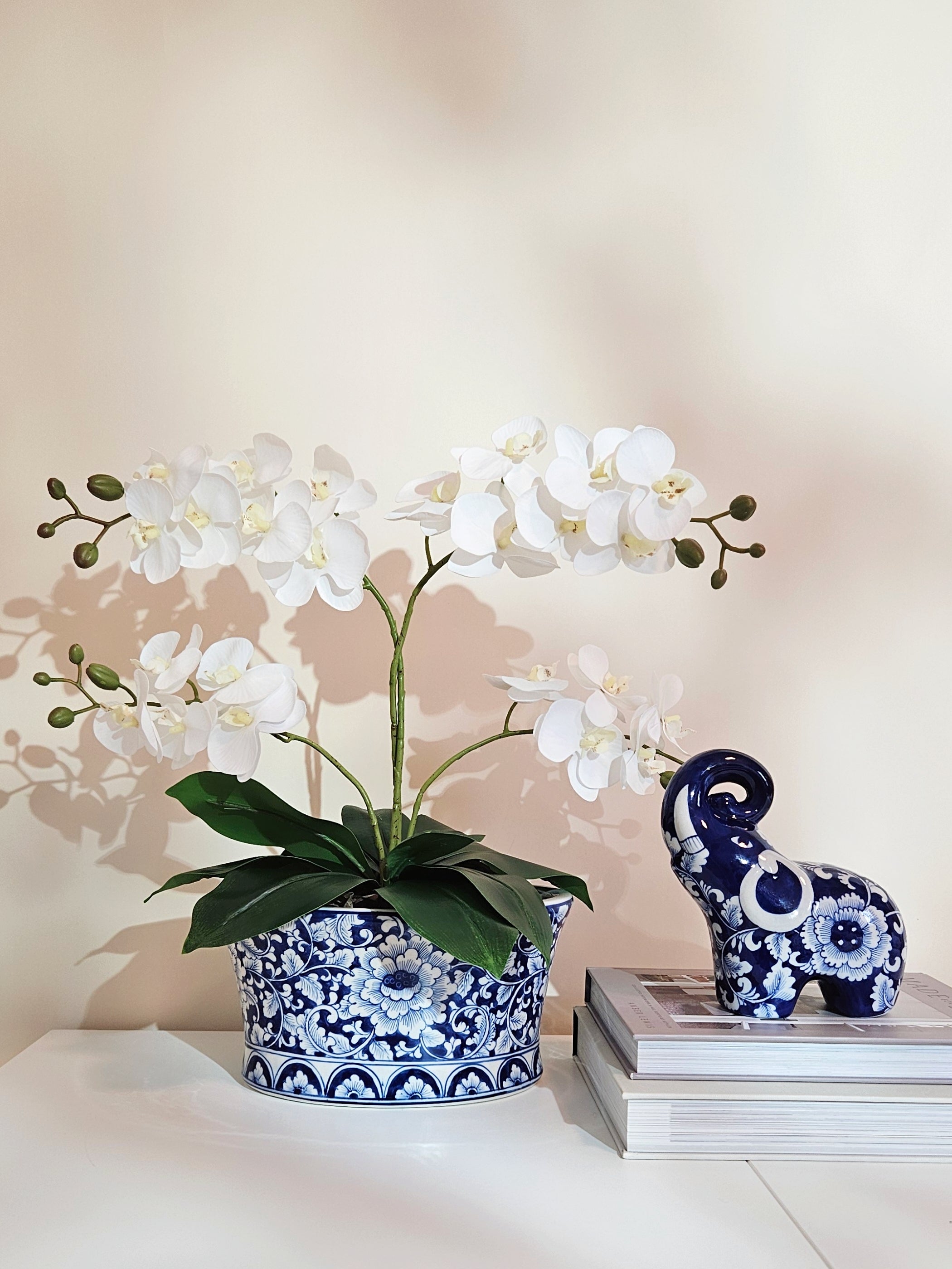 White artificial orchid flowers in a blue and white patterned ceramic planter pot, styled next to a matching blue and white elephant figurine on a stack of books against a warm beige wall.