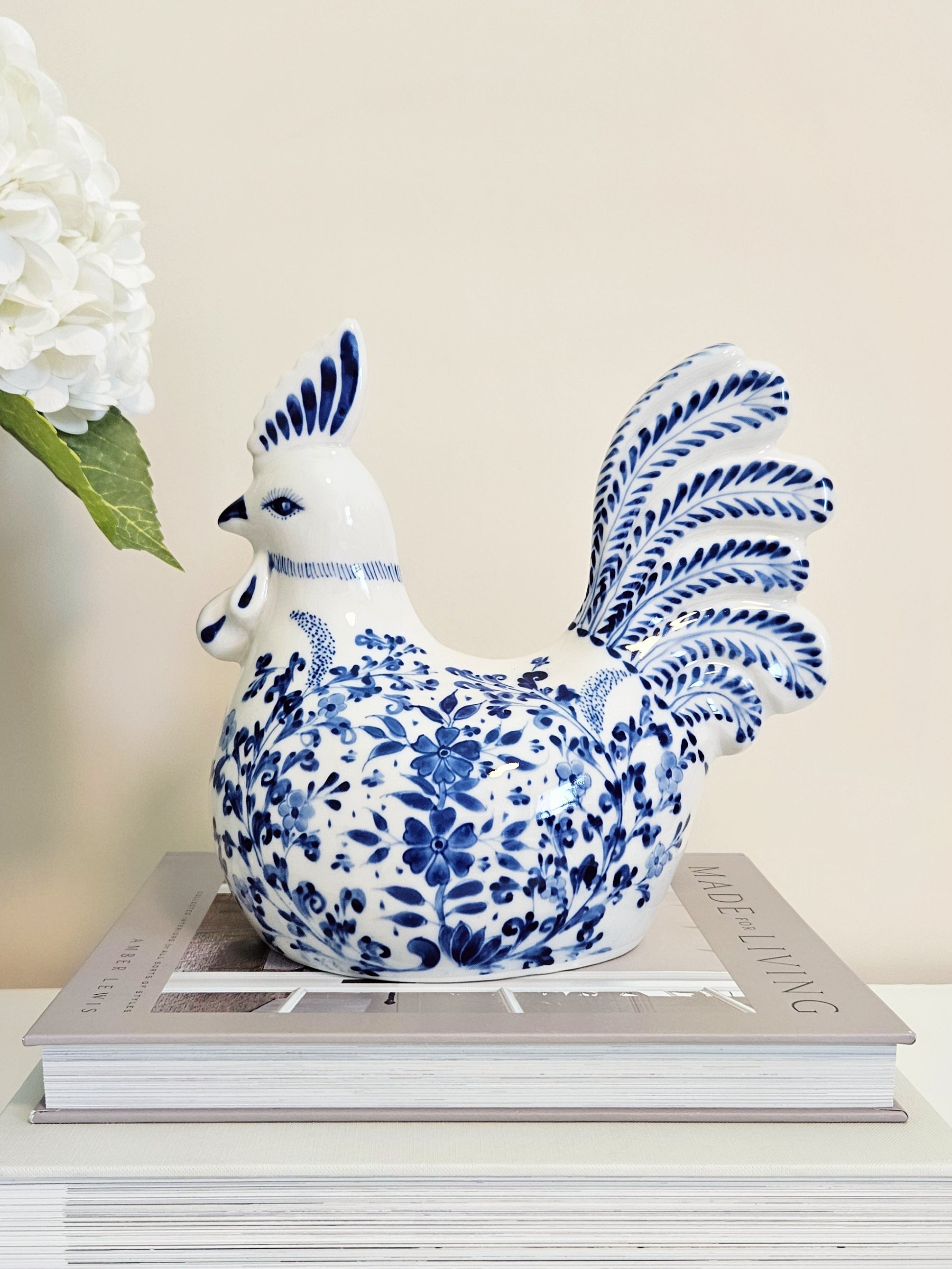 A ceramic rooster figurine in a classic blue and white floral pattern, displayed on top of a stack of books with a white flower in the background.