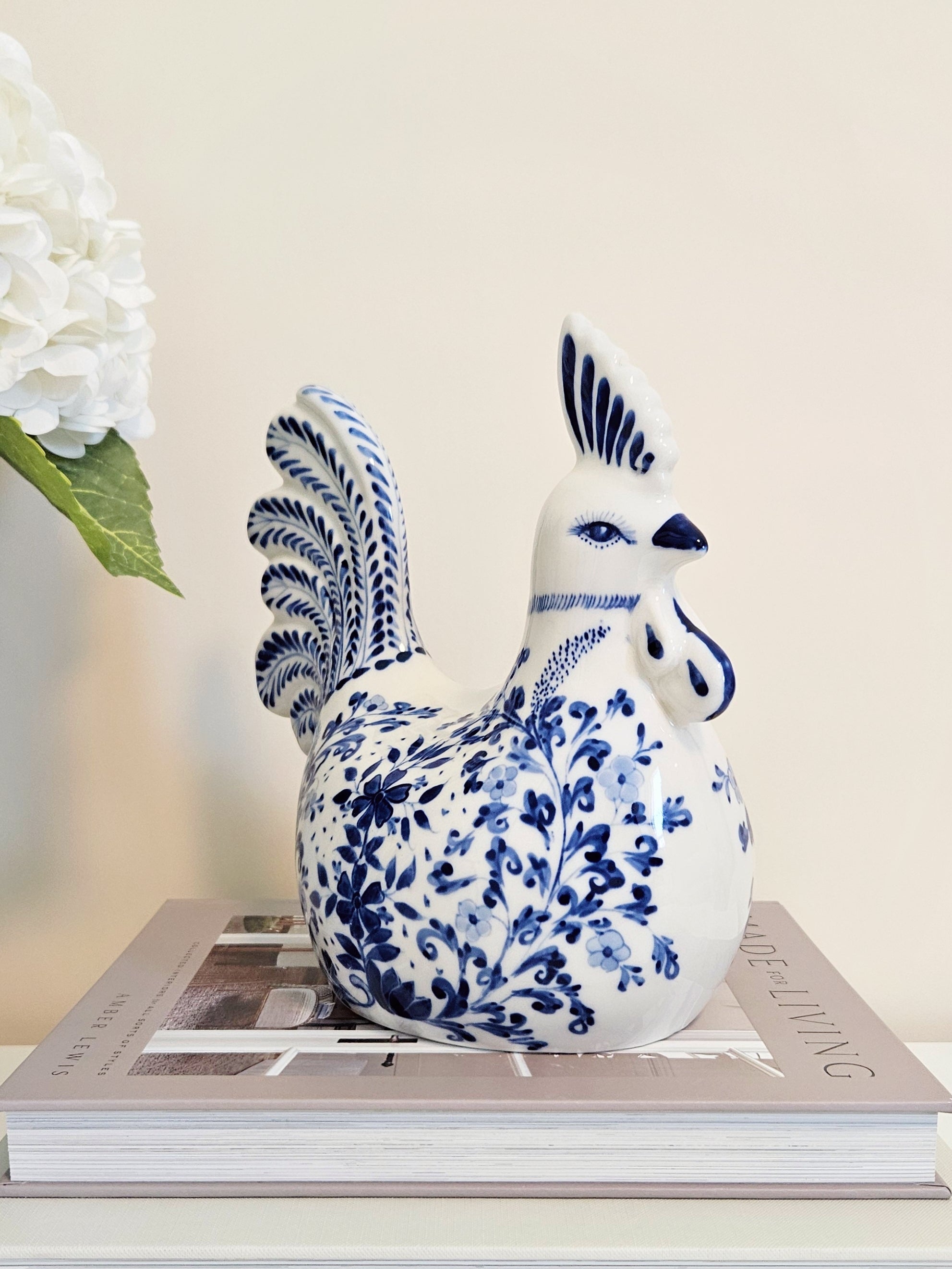 A ceramic rooster figurine with blue floral patterns on a white glaze, displayed on top of a book with a white flower in the background.