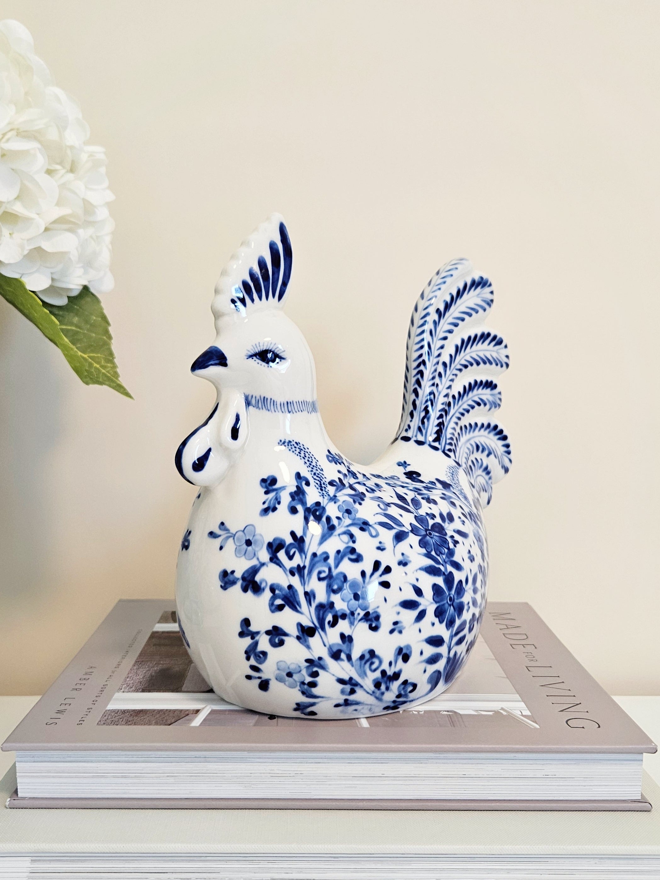 A ceramic rooster figurine with blue floral patterns on a white glaze, chinoiserie style , displayed on top of a book with a white flower in the background.