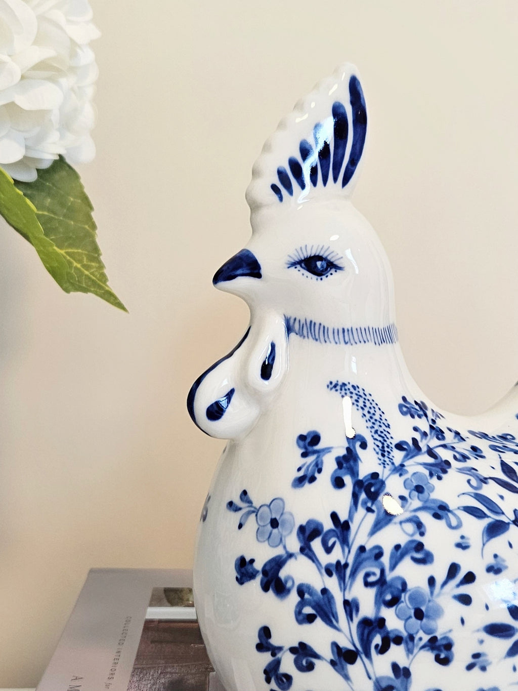 A white ceramic decorative rooster figurine with blue floral and bird patterns, shown in a close-up studio-style shot against a light beige background with a white flower and green leaf partially visible in the upper left.
