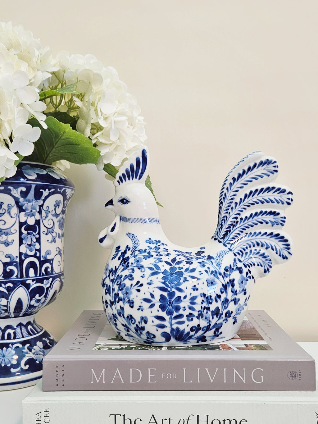 A blue and white porcelain rooster figurine sitting on top of a gray book, next to a matching blue and white floral vase with white flowers, against a warm beige background.