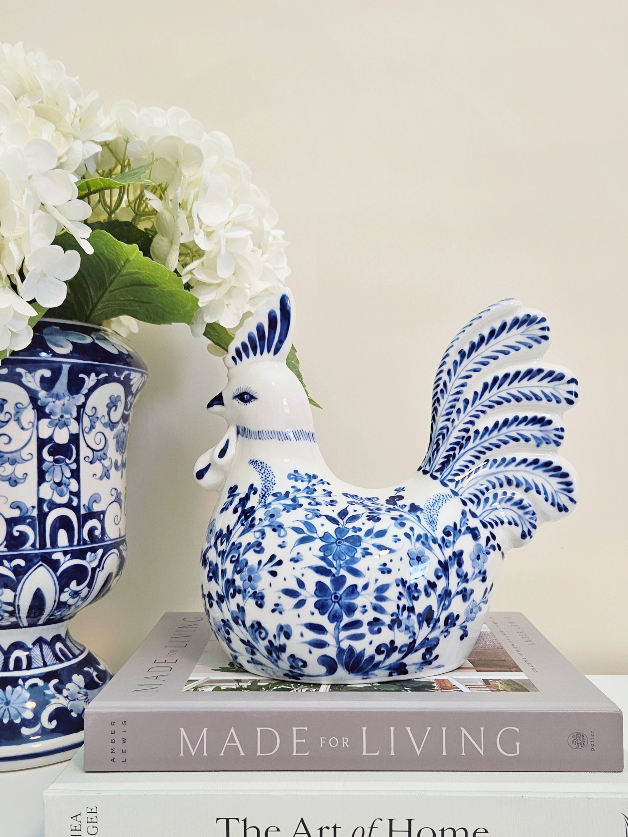A blue and white porcelain rooster figurine sitting on top of a gray book, next to a matching blue and white floral vase with white flowers, against a warm beige background.