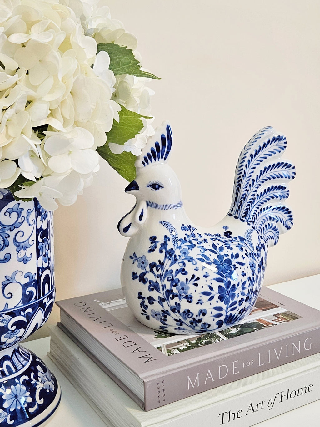 A blue and white porcelain rooster figurine decorated with floral patterns, displayed on a stack of books next to a matching blue and white patterned vase with white flowers.