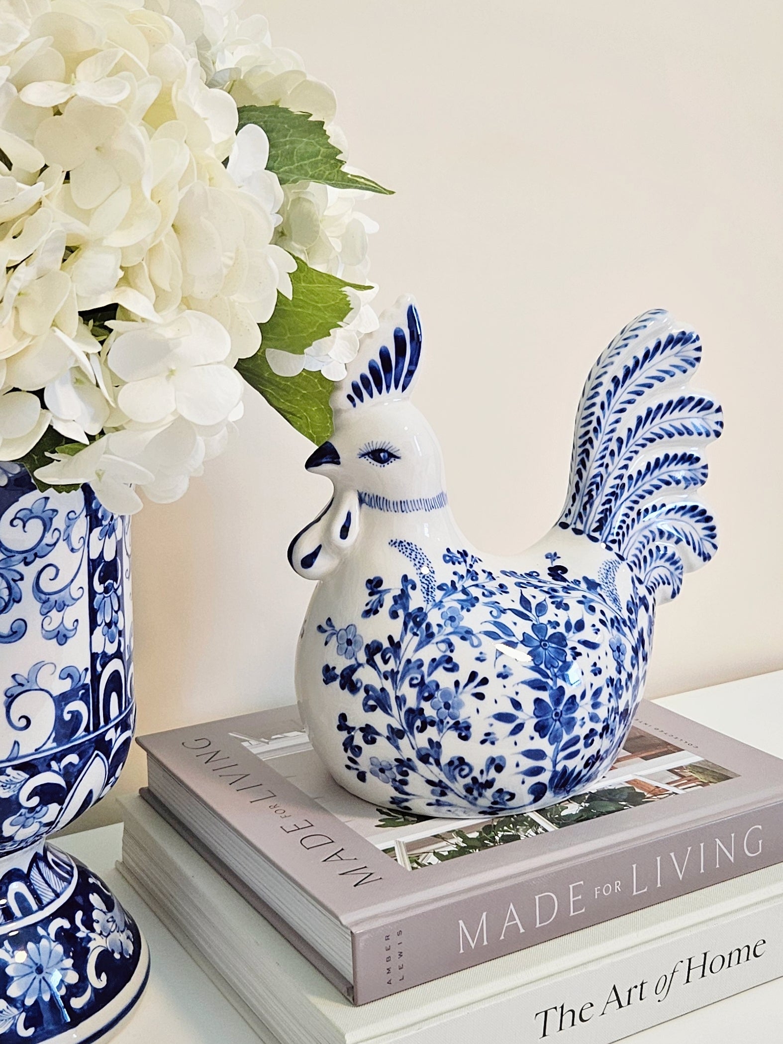 A blue and white porcelain rooster figurine decorated with floral patterns, displayed on a stack of books next to a matching blue and white patterned vase with white flowers.