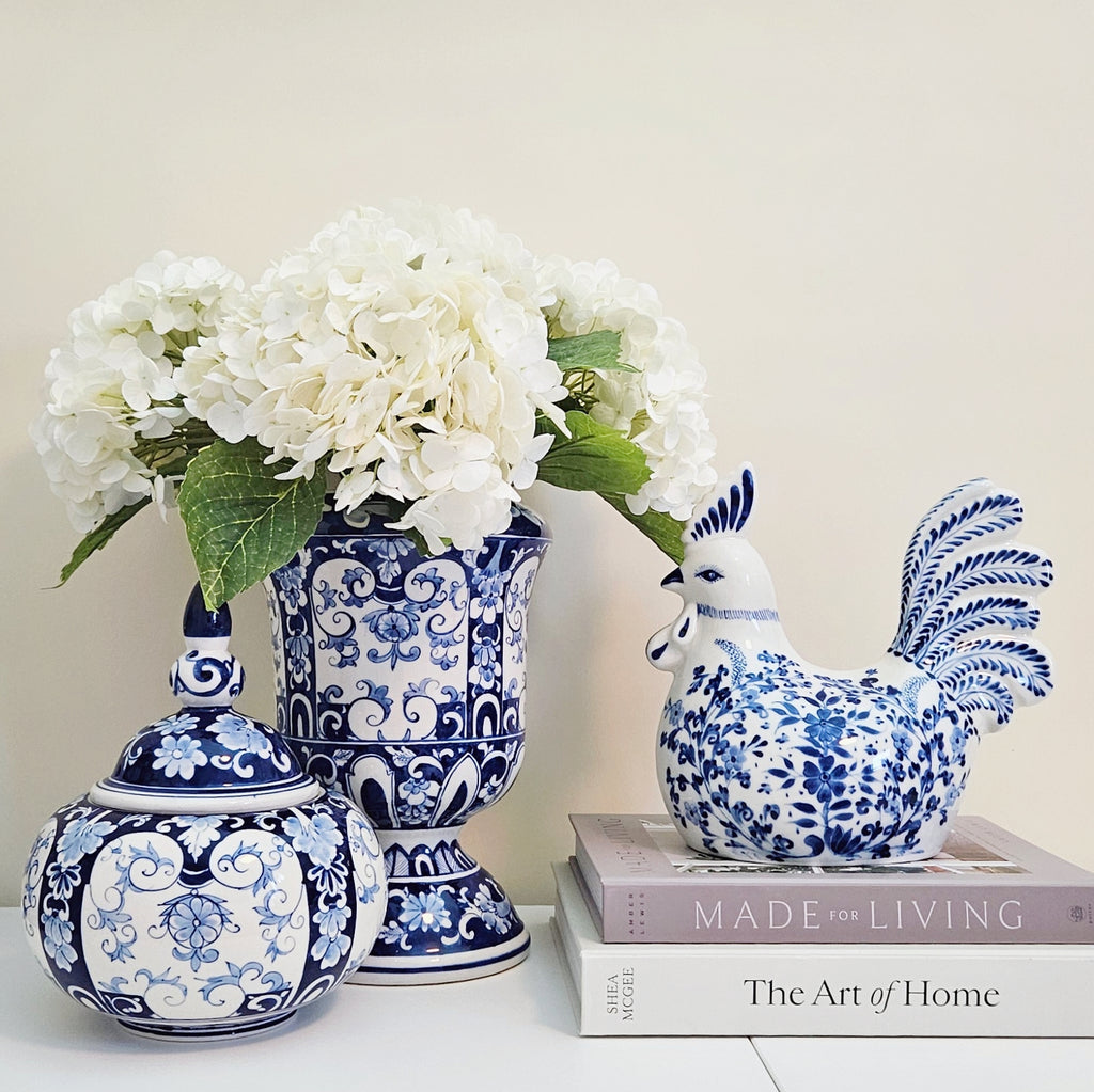 A styled arrangement of blue and white porcelain home decor on a white surface against a warm beige wall. The set includes a lidded vase with a floral rooster figurine, a tall pedestal vase filled with white hydrangeas, and a small lidded urn.