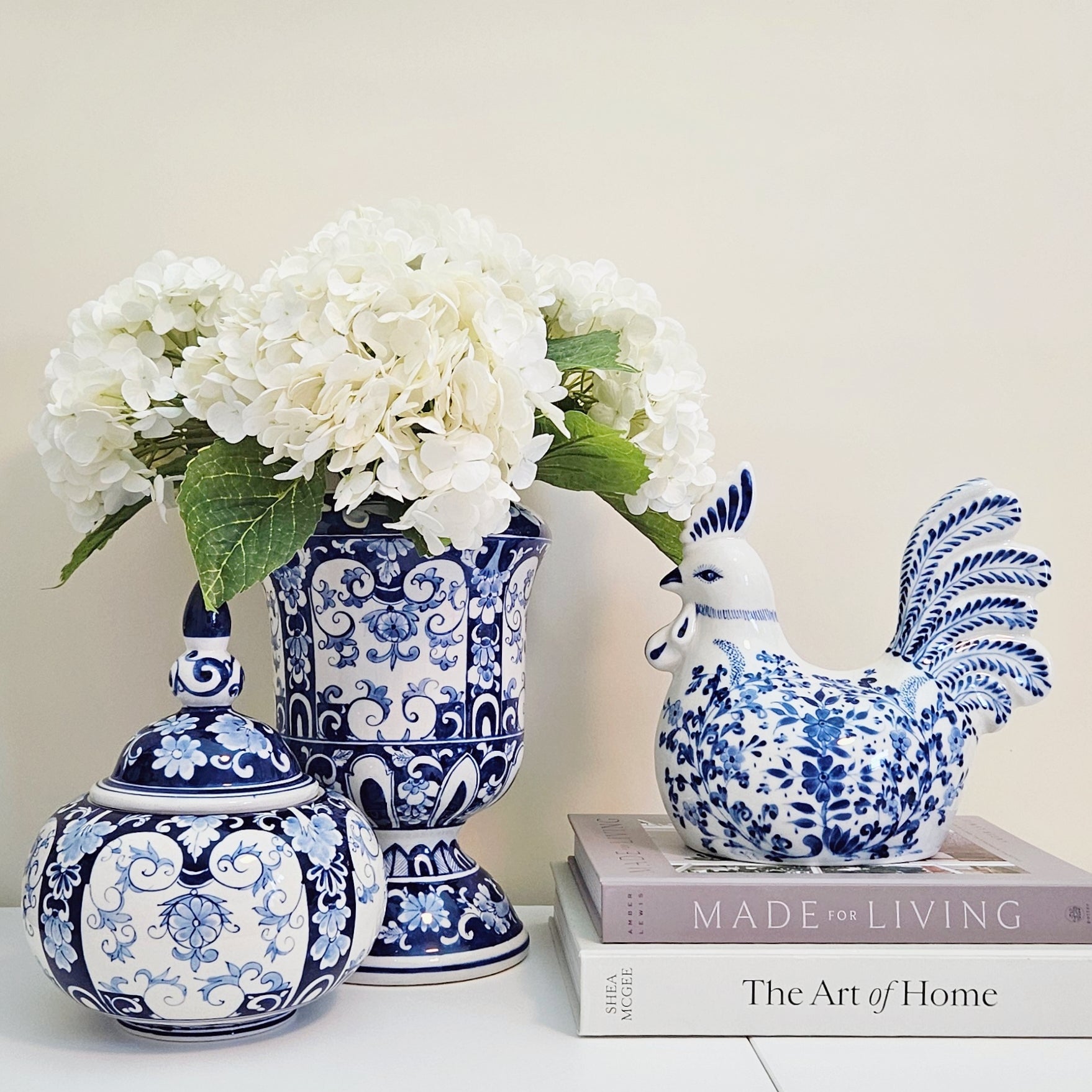 A styled arrangement of blue and white porcelain home decor on a white surface against a warm beige wall. The set includes a lidded vase with a floral rooster figurine, a tall pedestal vase filled with white hydrangeas, and a small lidded urn.