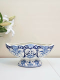 A blue and white porcelain scalloped footed planter or pedestal bowl with a decorative floral pattern, displayed on a white surface against a beige wall with white lilies in the background.