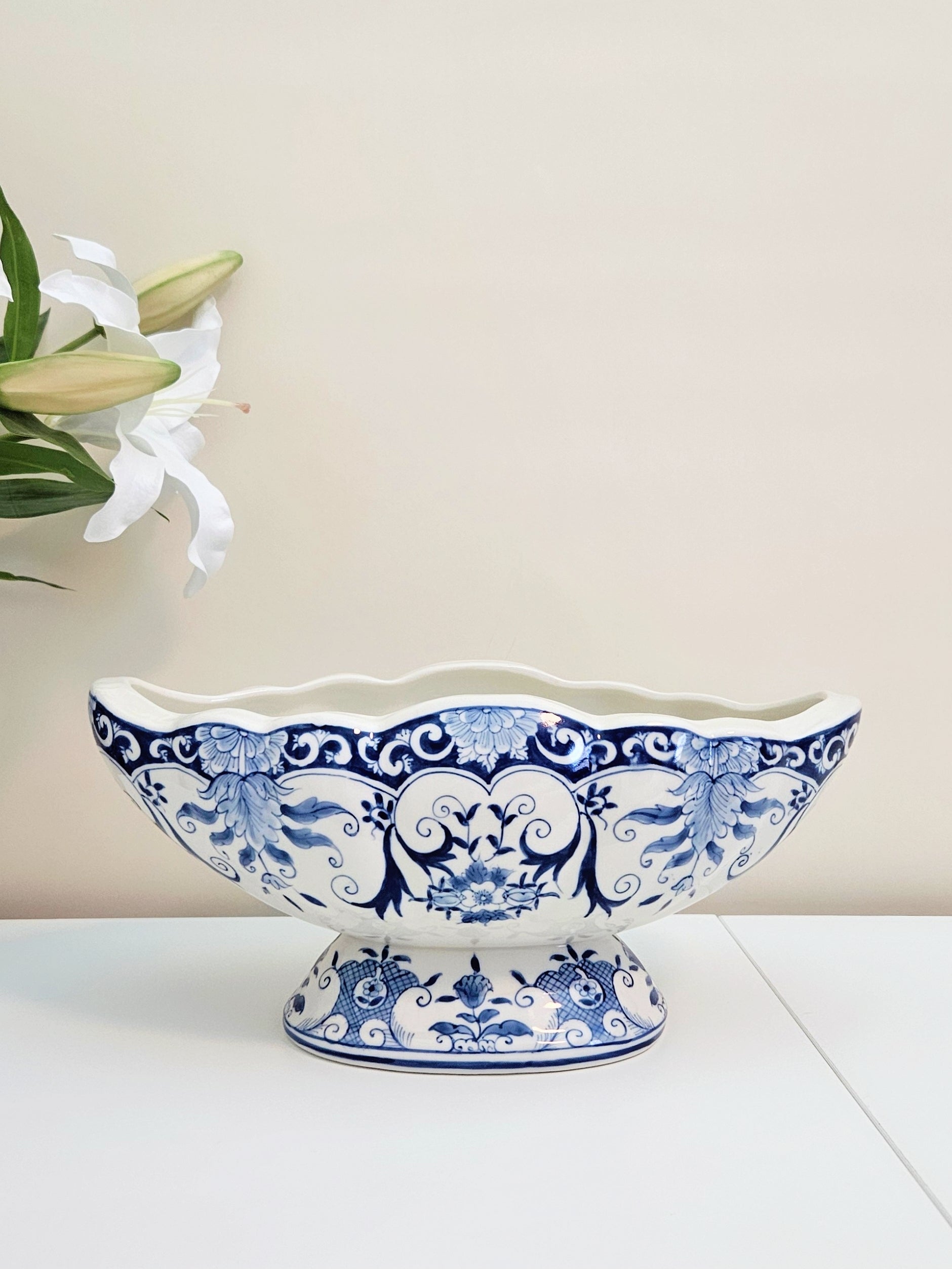A blue and white porcelain scalloped footed planter or pedestal bowl with a decorative floral pattern, displayed on a white surface against a beige wall with white lilies in the background.