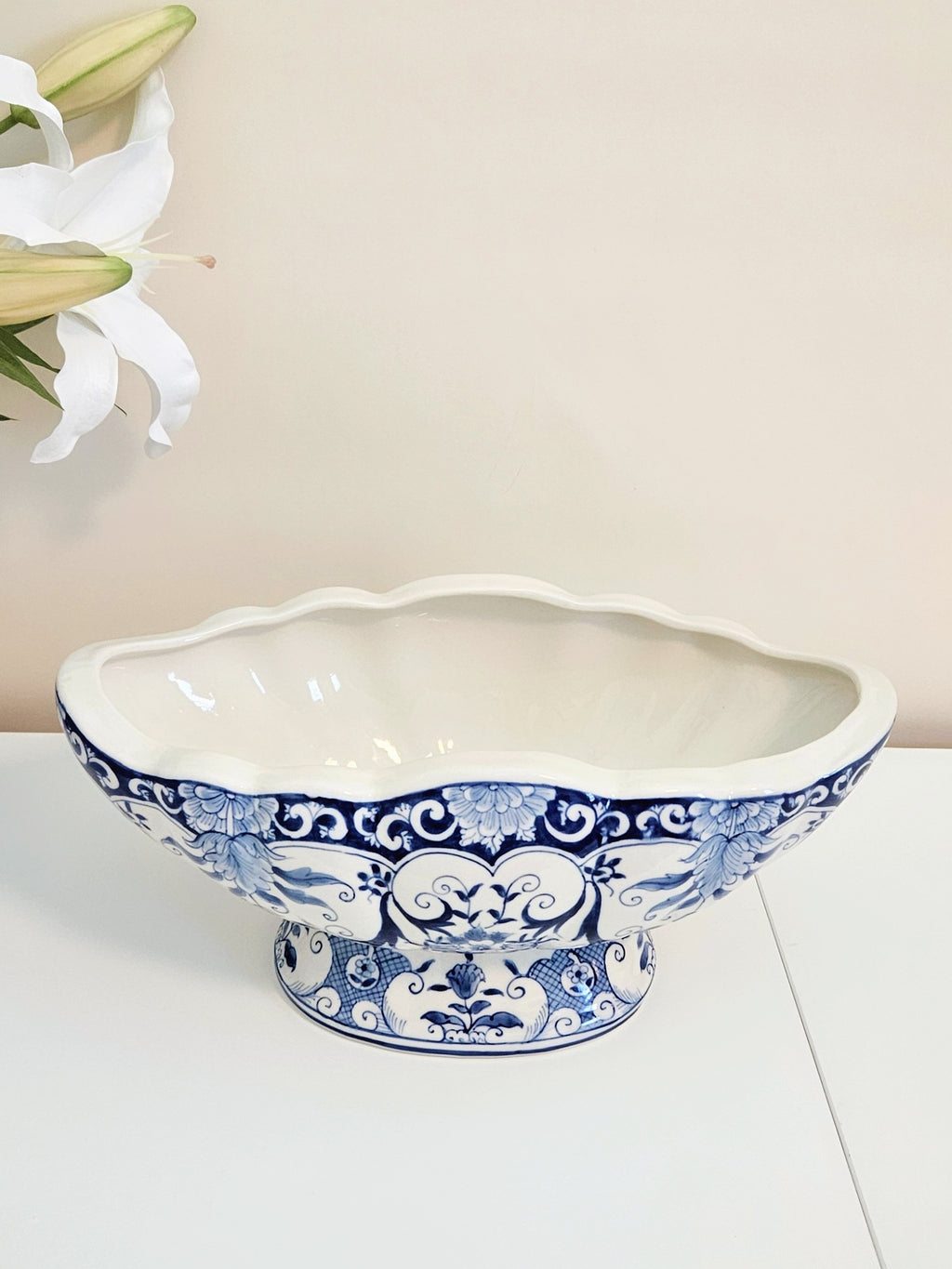 A blue and white porcelain bowl or planter with a scalloped rim and ornate floral patterns, displayed on a white surface against a light beige background with a white lily flower in the upper left corner.