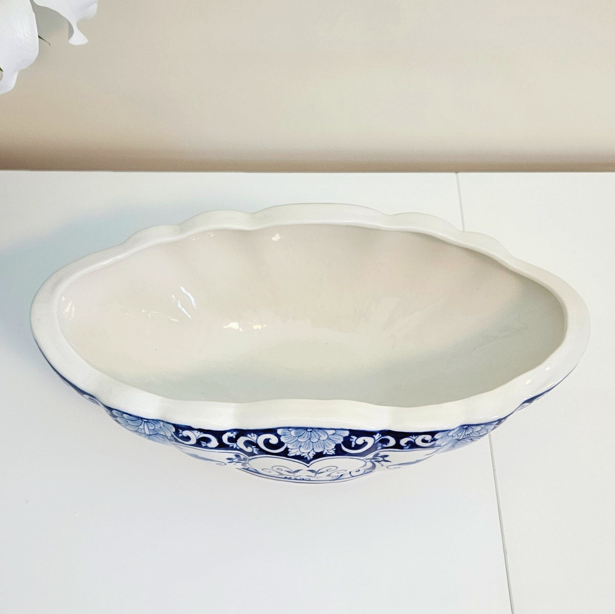 An oval ceramic bowl or planter with a scalloped rim, featuring a glossy white interior and a blue and white floral scroll pattern around the lower exterior, shown on a white surface against a beige wall.