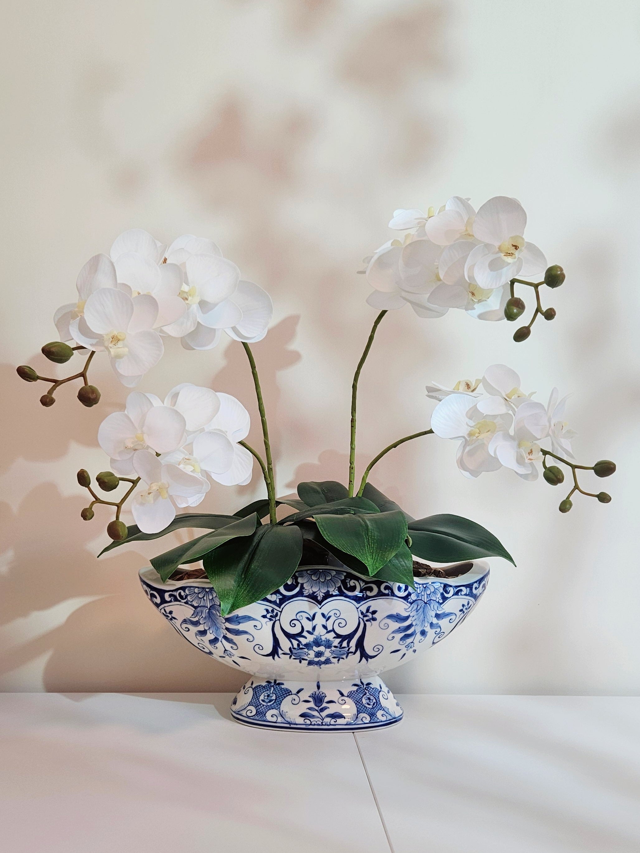 White artificial Phalaenopsis orchid flowers with green stems and leaves, arranged in a blue and white scalloped porcelain footed planter or pedestal bowl on a light surface against a softly lit neutral background.
