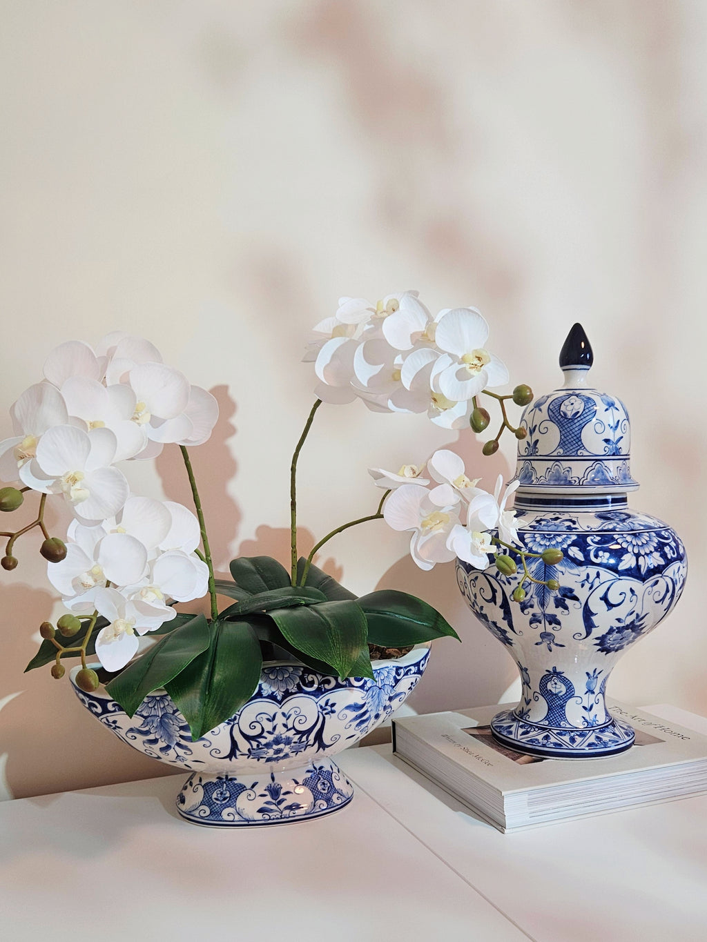 A blue and white porcelain orchid planter with white artificial orchid flowers and green leaves, displayed next to a matching blue and white lidded ginger jar on a stack of books against a soft beige background.