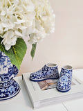 A pair of small blue and white floral-patterned shoe figurines displayed on top of a magazine next to a large white hydrangea arrangement in a blue-and-white vase against a soft beige wall.