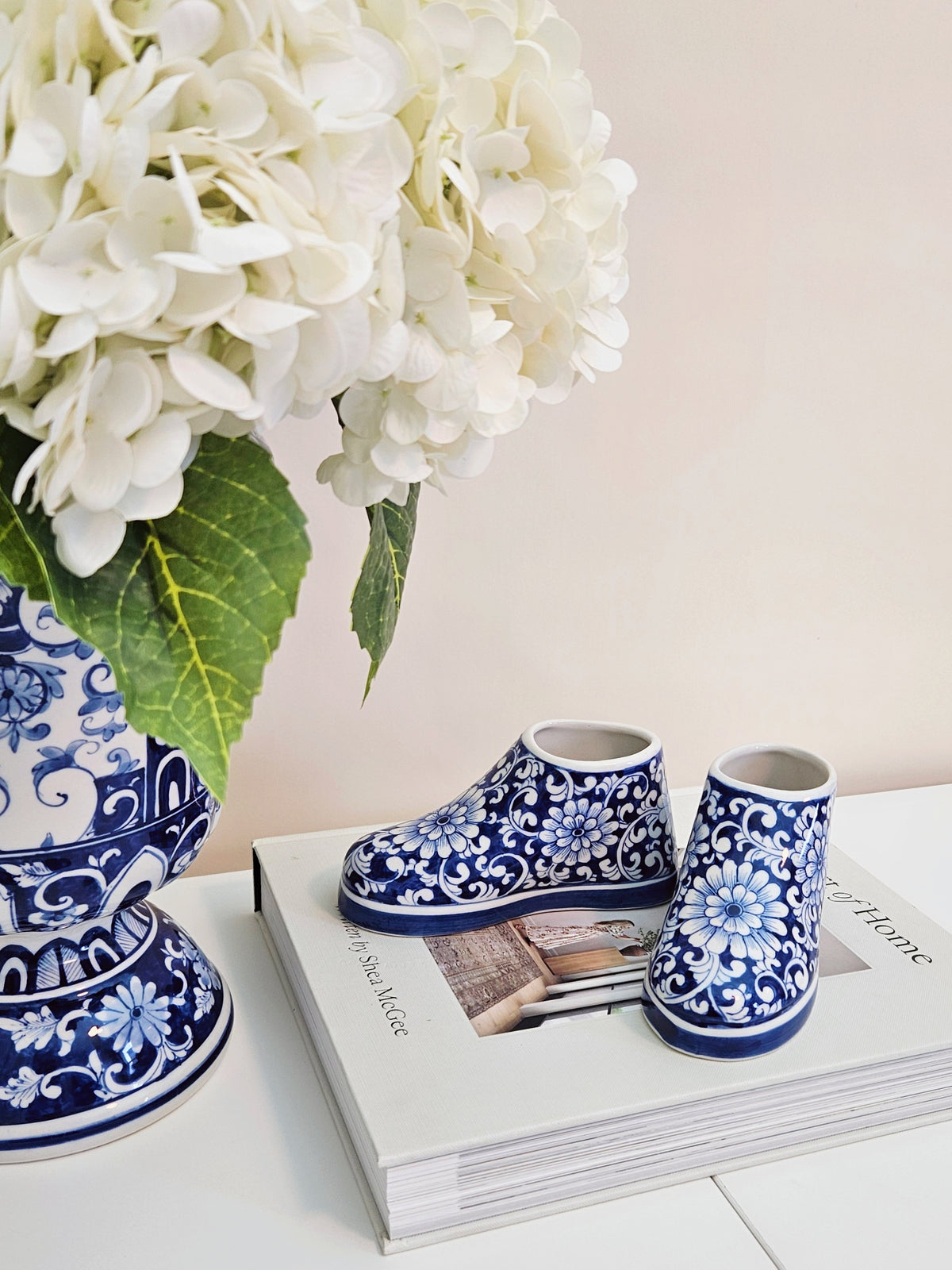 A pair of small blue and white floral-patterned shoe figurines displayed on top of a magazine next to a large white hydrangea arrangement in a blue-and-white vase against a soft beige wall.