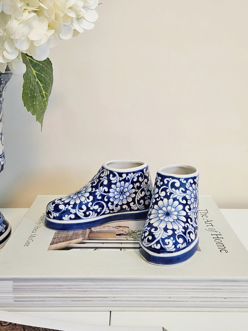A pair of blue and white floral-patterned ceramic booties resting on a stack of books, with a vase of white flowers in the background against a light beige wall.