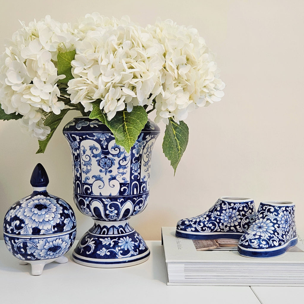 Blue and white porcelain-style decorative set including a tall urn-shaped vase with white flowers, a small lidded jar, and a pair of bootie-shaped figurines, arranged on a white surface against a light beige background.