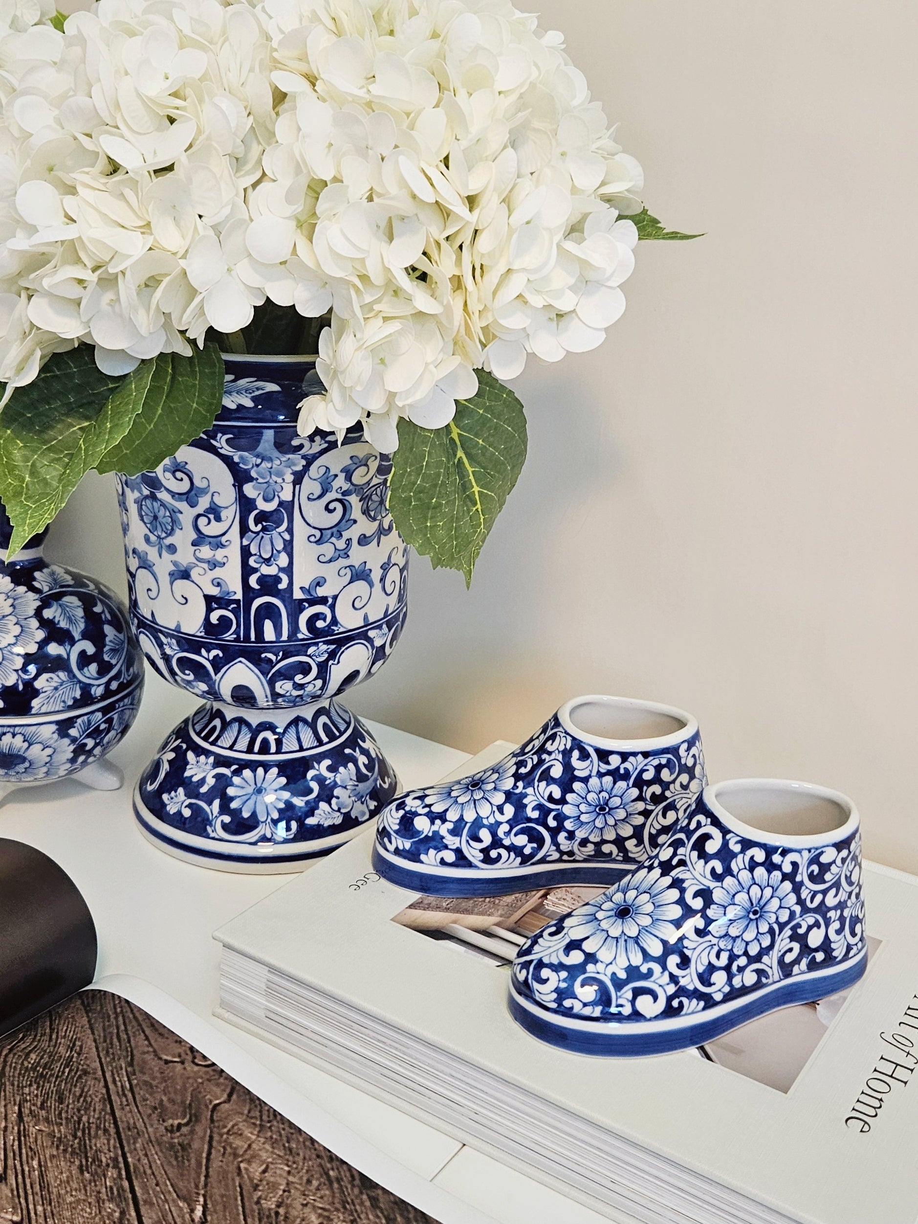 A pair of small  porcelain blue and white floral-patterned booties styled on a white book next to a matching blue and white ceramic vase filled with white flowers.