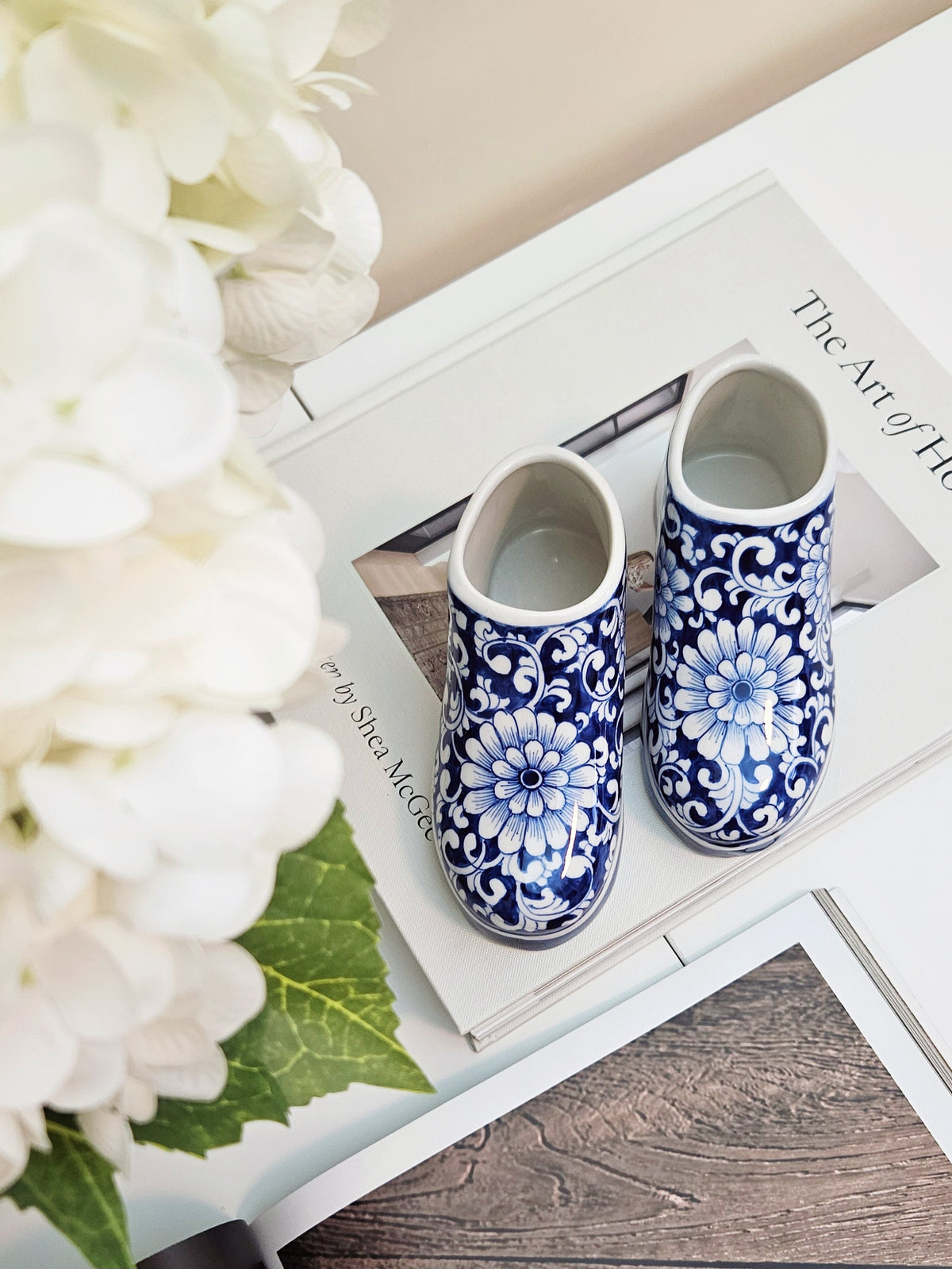 A pair of small ceramic blue and white floral ceramic shoes or dollhouse figures placed on top of open book pages, with white flowers and a wooden surface around them.