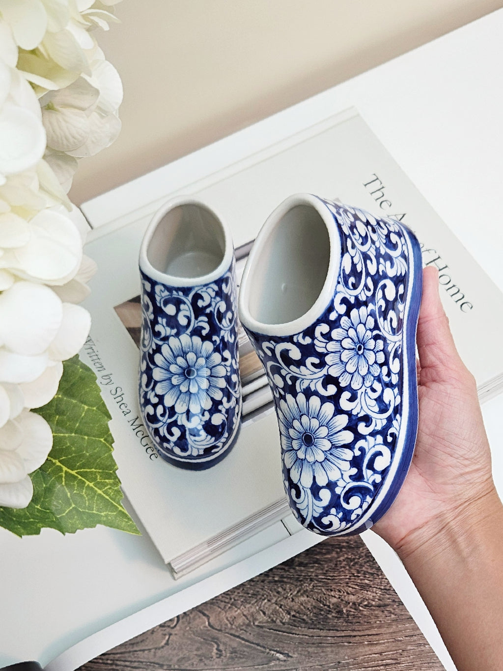 A pair of ceramic blue and white floral-patterned slip-on shoes, with one held in a hand over a white book and white flowers.