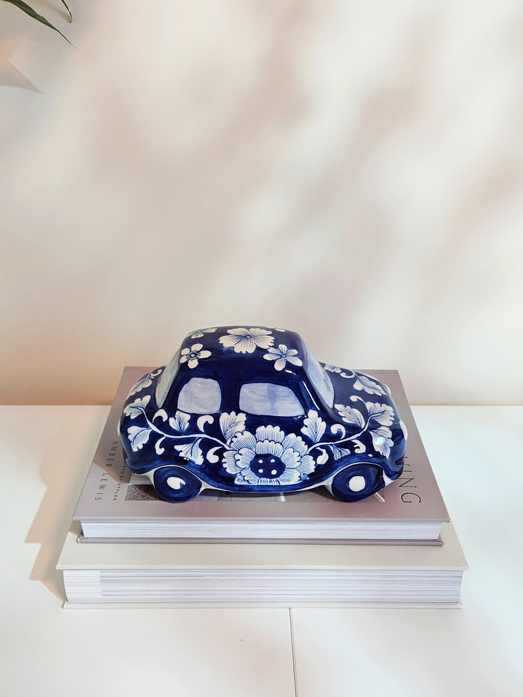 A glossy ceramic car figurine decorated with blue and white floral patterns, chinoiserie style, resting on a stack of two books against a softly lit beige background.